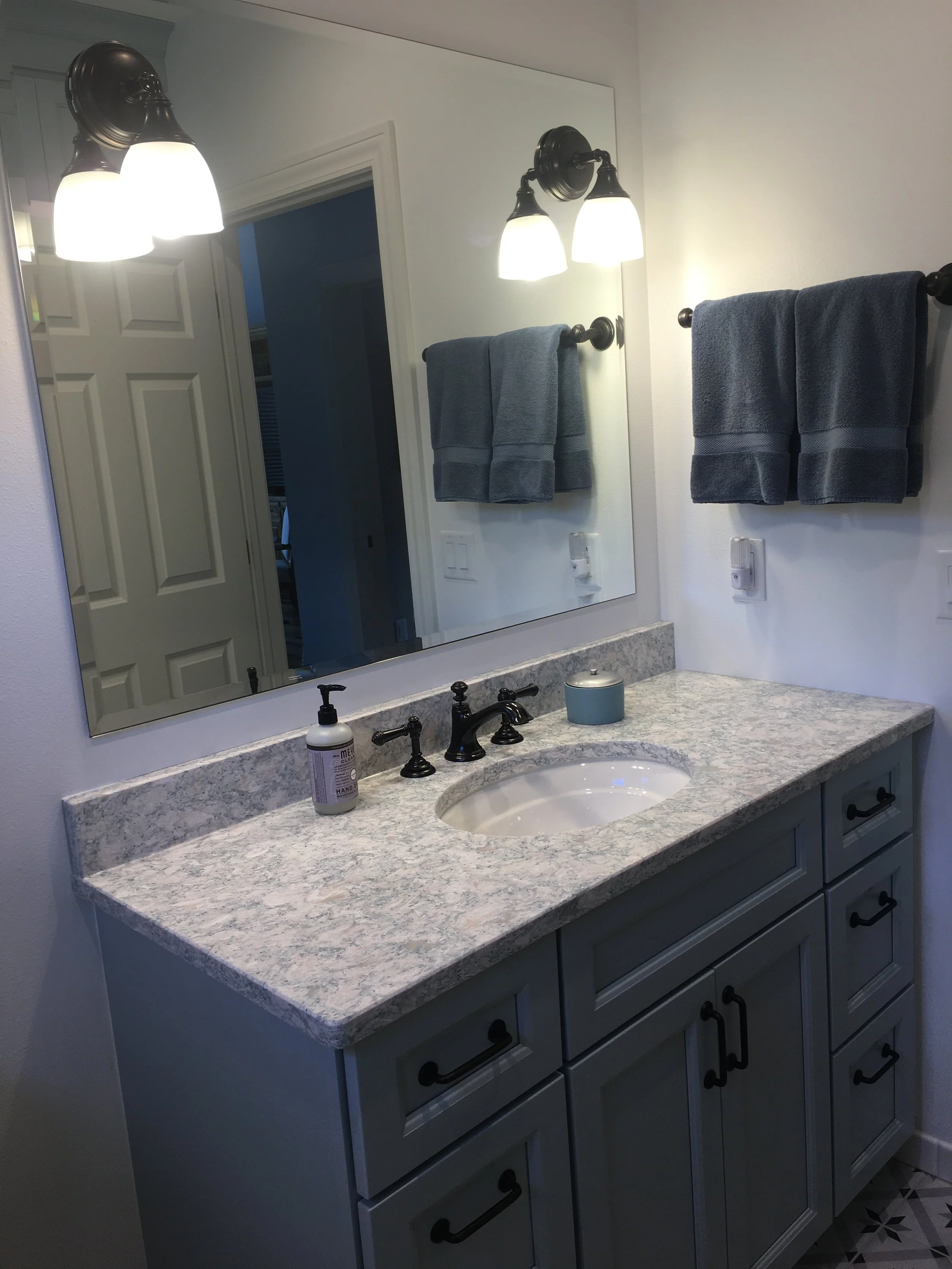 Bathroom vanity with a granite countertop, oval sink, soap dispenser, and a small container. Two gray towels hang on a towel bar, with a large mirror above reflecting part of the room. Two light fixtures with downward-facing frosted glass shades are 