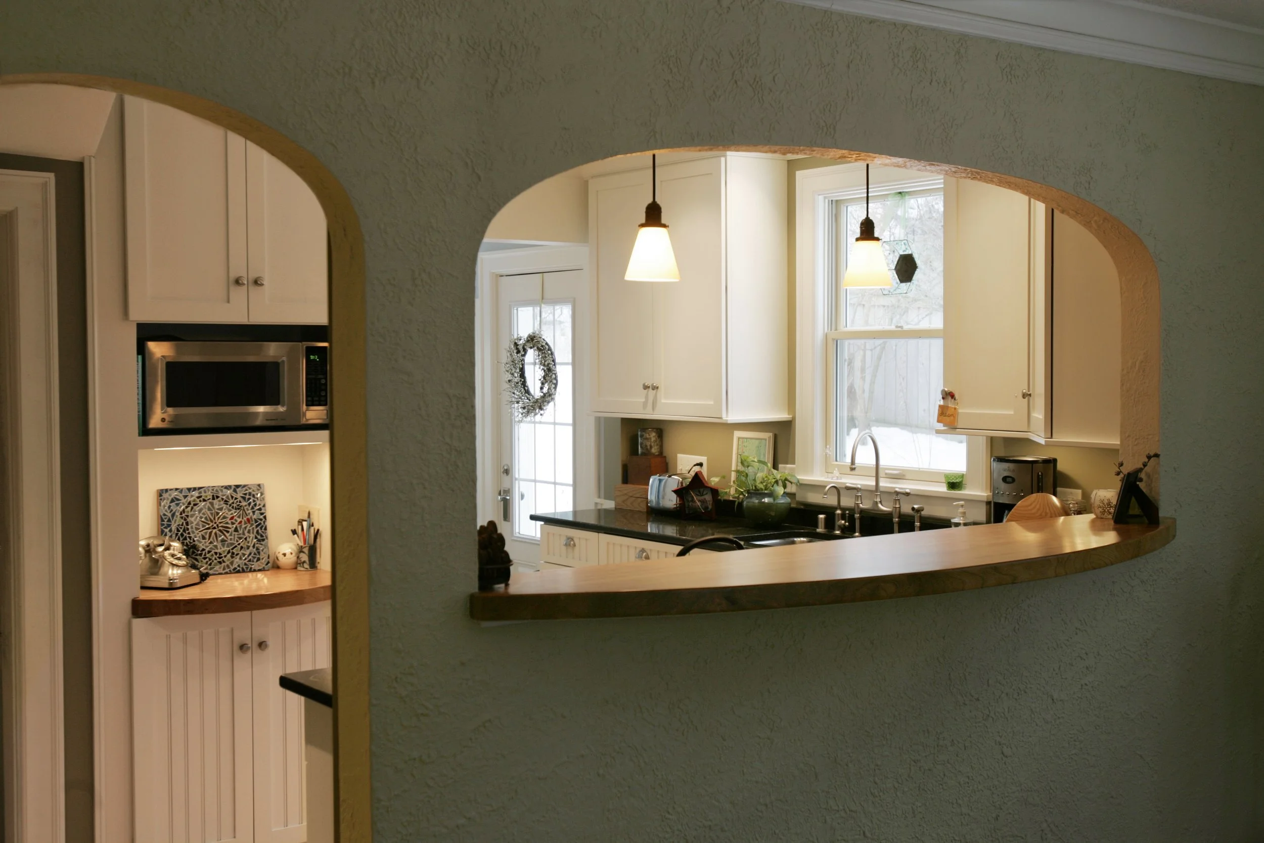 View of a kitchen through a curved counter window with white cabinets, black countertop, and a window with a view outside.