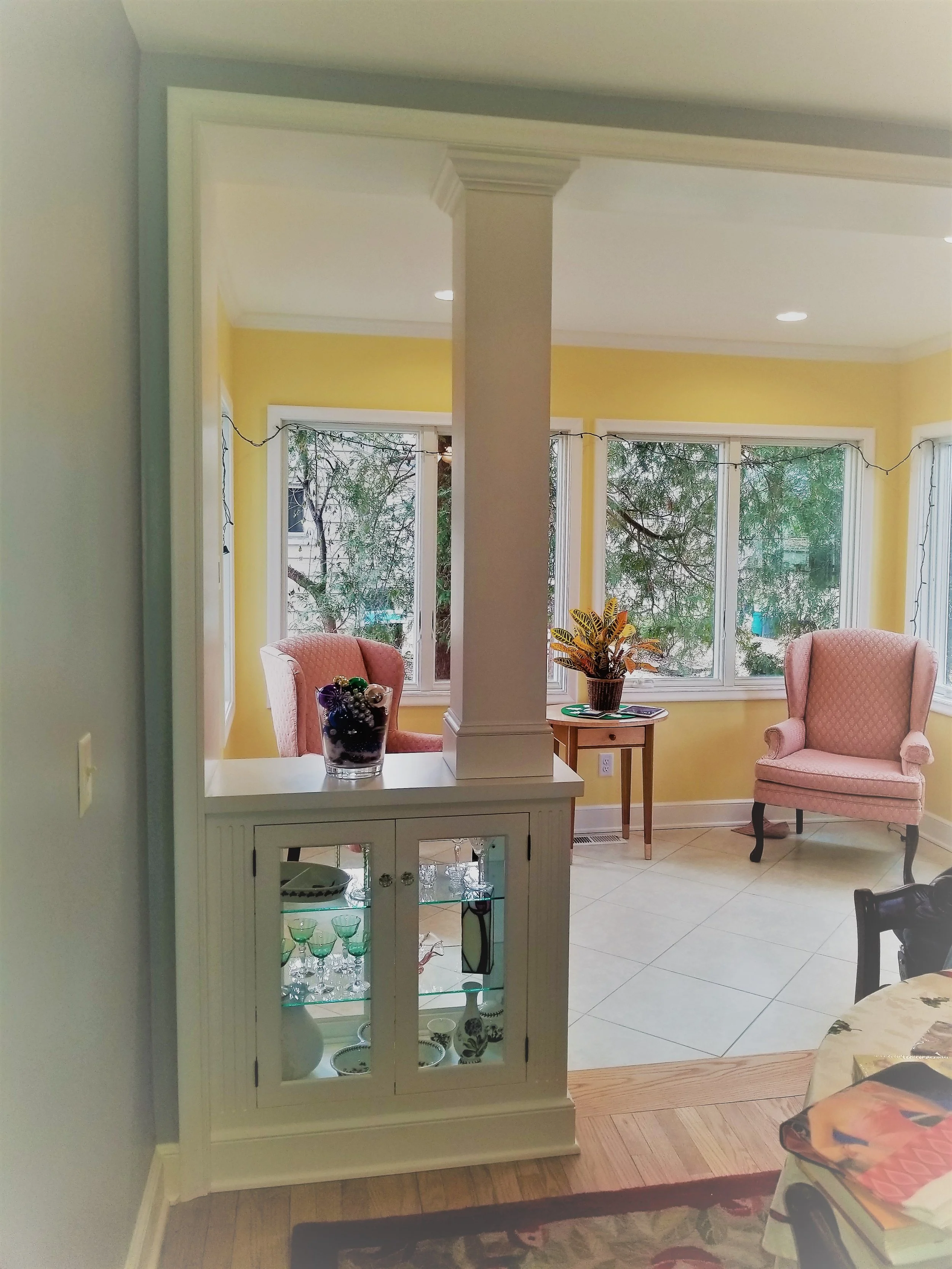 Living room with yellow walls, large windows, pink armchairs, a small wooden table with a potted plant, decorative string lights, and a white display cabinet with glass doors containing glassware.