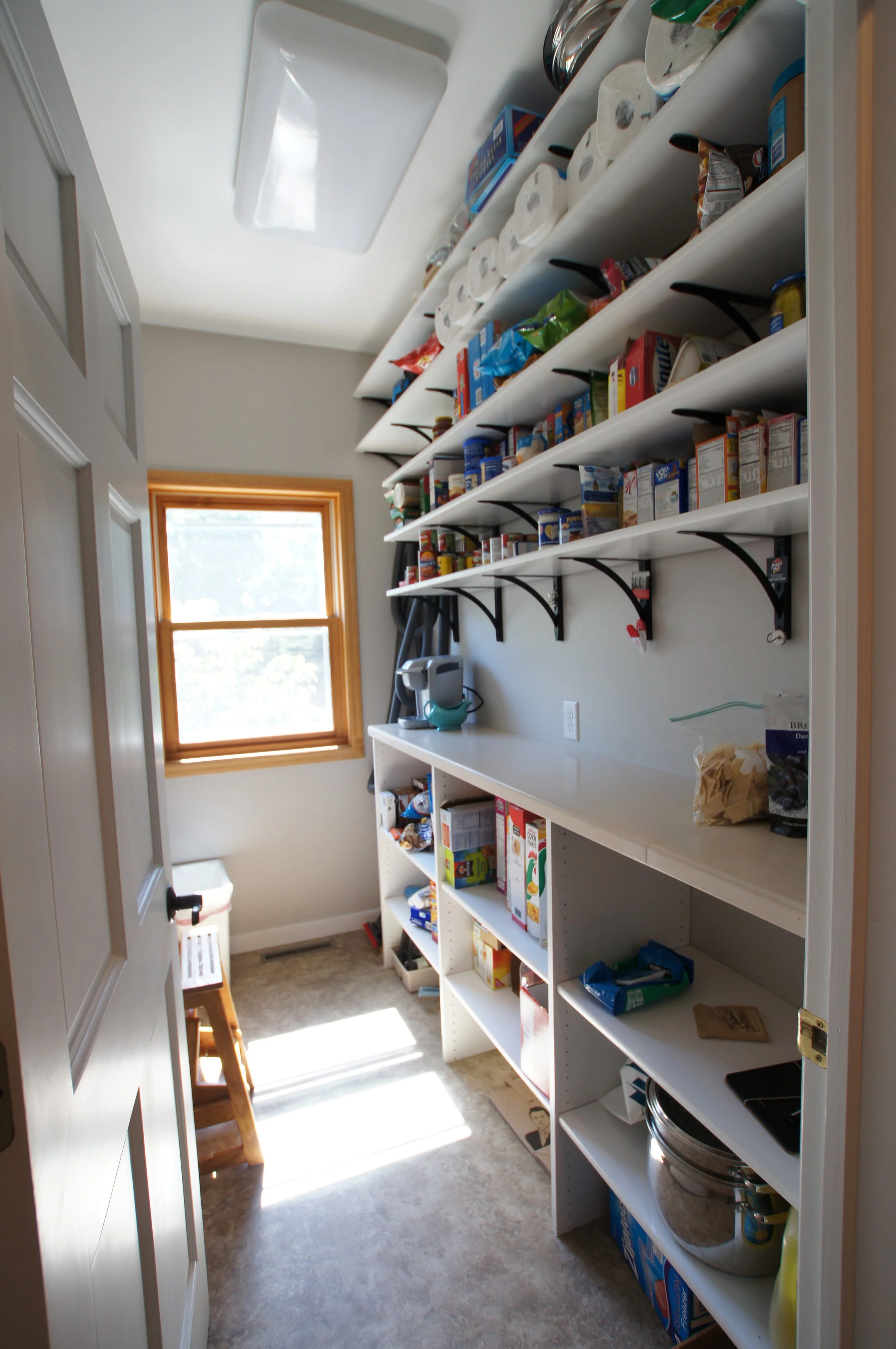 A small pantry with shelves filled with canned goods, snacks, and paper towels, a window letting in sunlight, and a small countertop with a coffee maker.