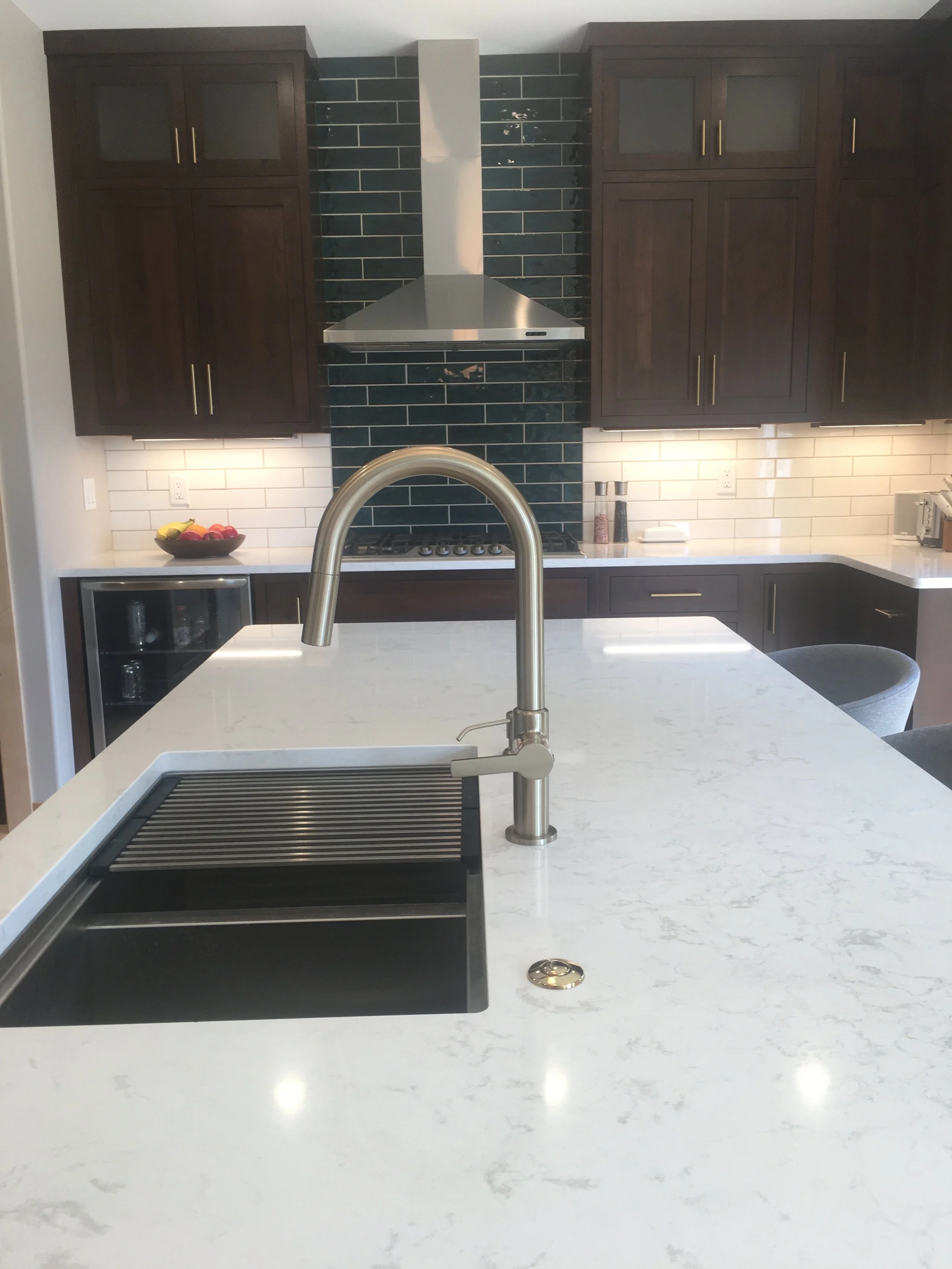 Modern kitchen with white marble island, stainless steel faucet, black sink, dark wood cabinets, white subway tile backsplash, dark blue tile behind stove, stainless steel range hood, and small appliances on the counter.
