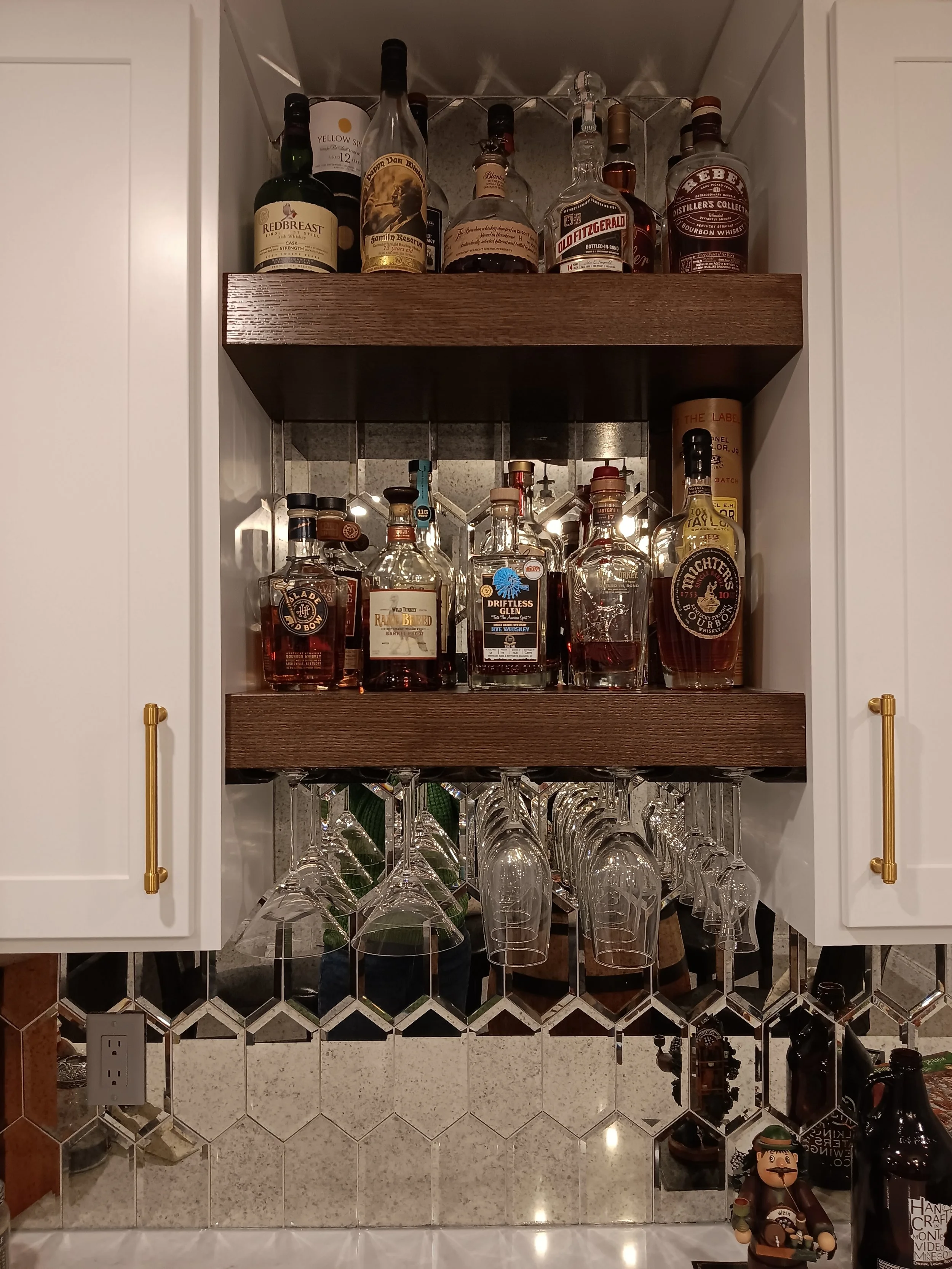 A kitchen cabinet with three shelves filled with various bottles of whiskey and liquor, including brands like Redbreast, Old Fitzgerald, and Rebel. The bottom shelf has hanging wine glasses, and the backsplash features a reflective tile pattern with 