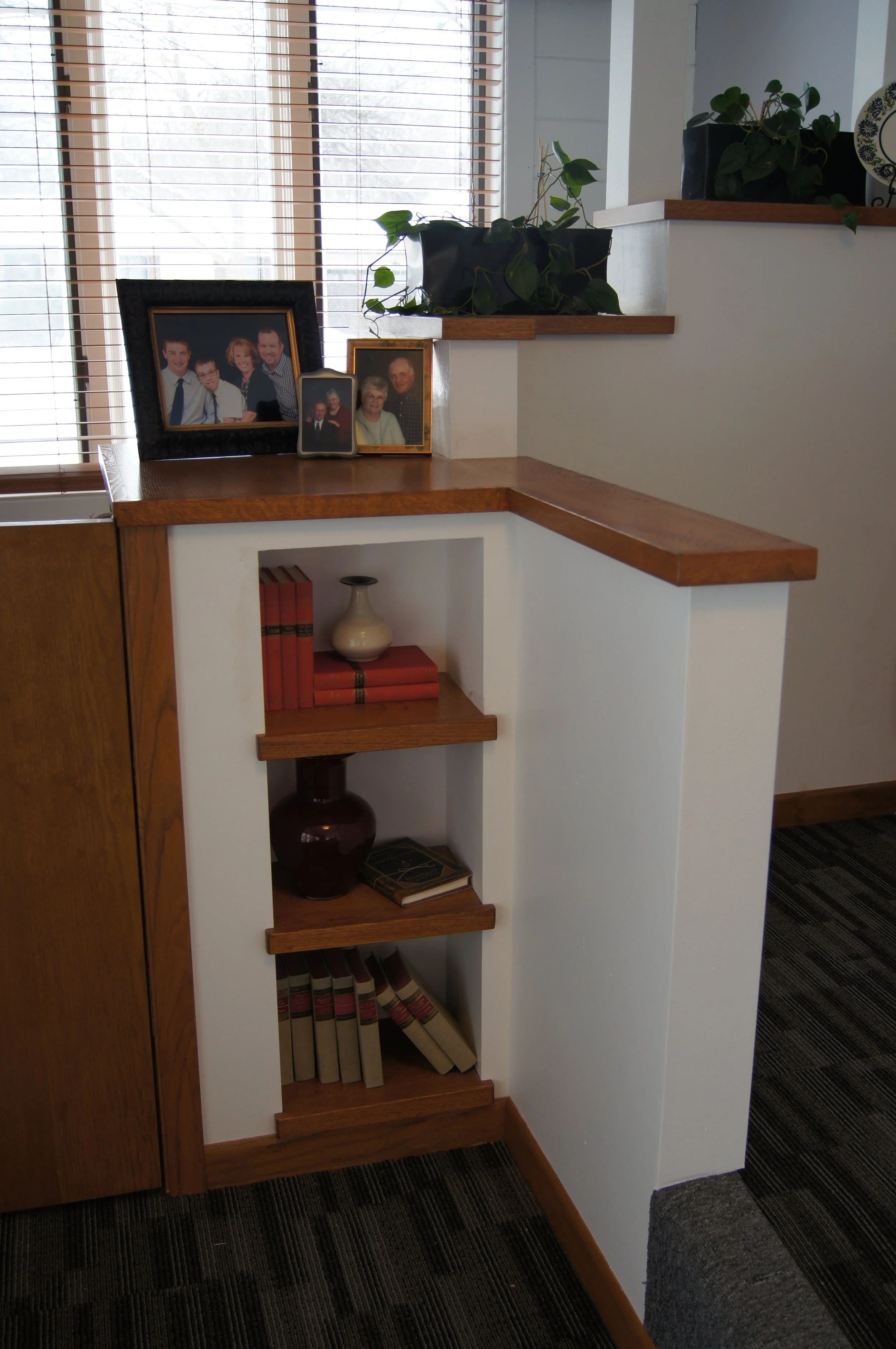 Interior of a home with a built-in corner bookshelf holding books, decorative vases, and framed photographs on a wooden ledge, with window blinds behind.