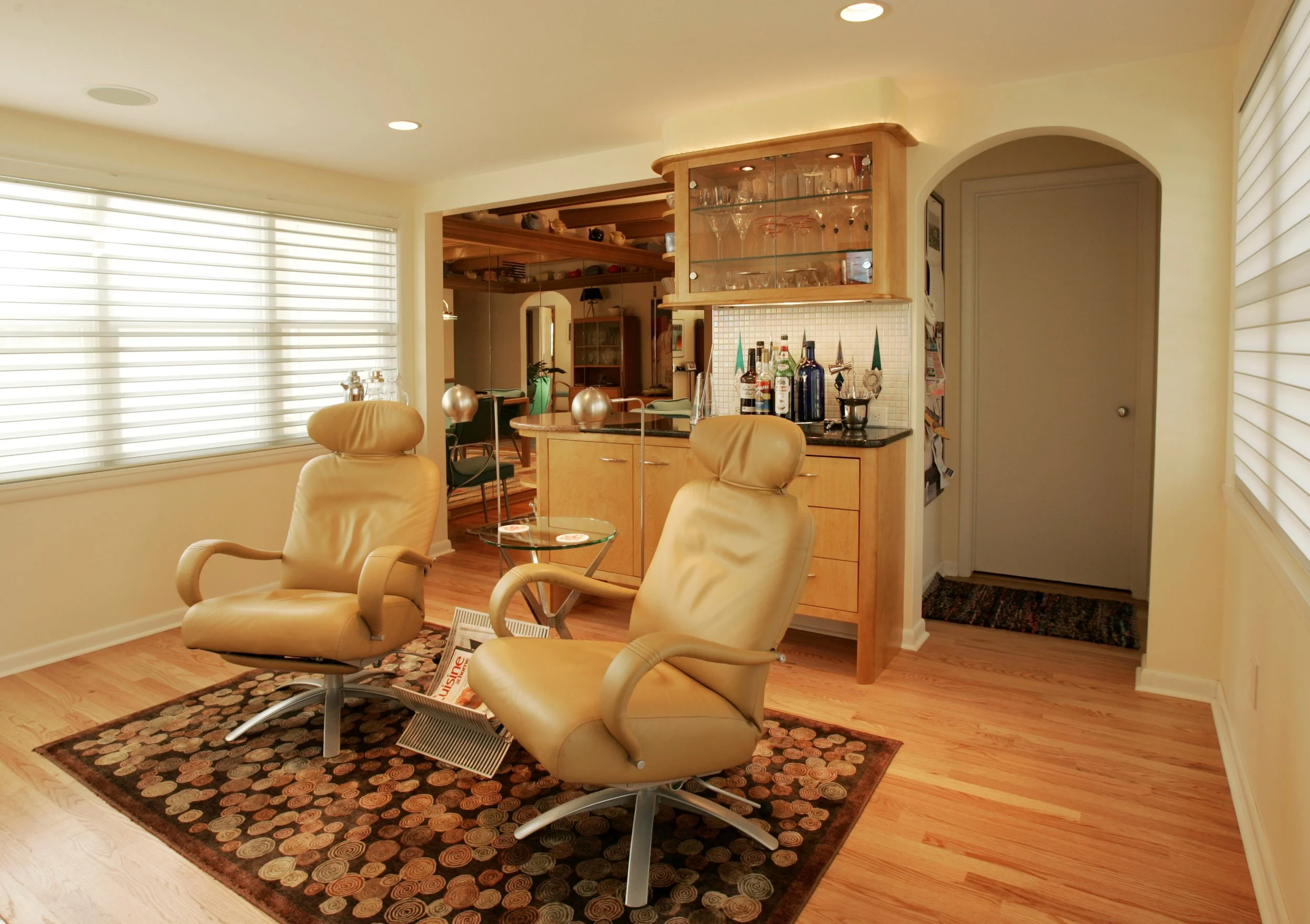 Living room with two tan leather chairs, a small glass table, a patterned area rug, large windows with blinds, and a bar area with bottles and glasses in the background.