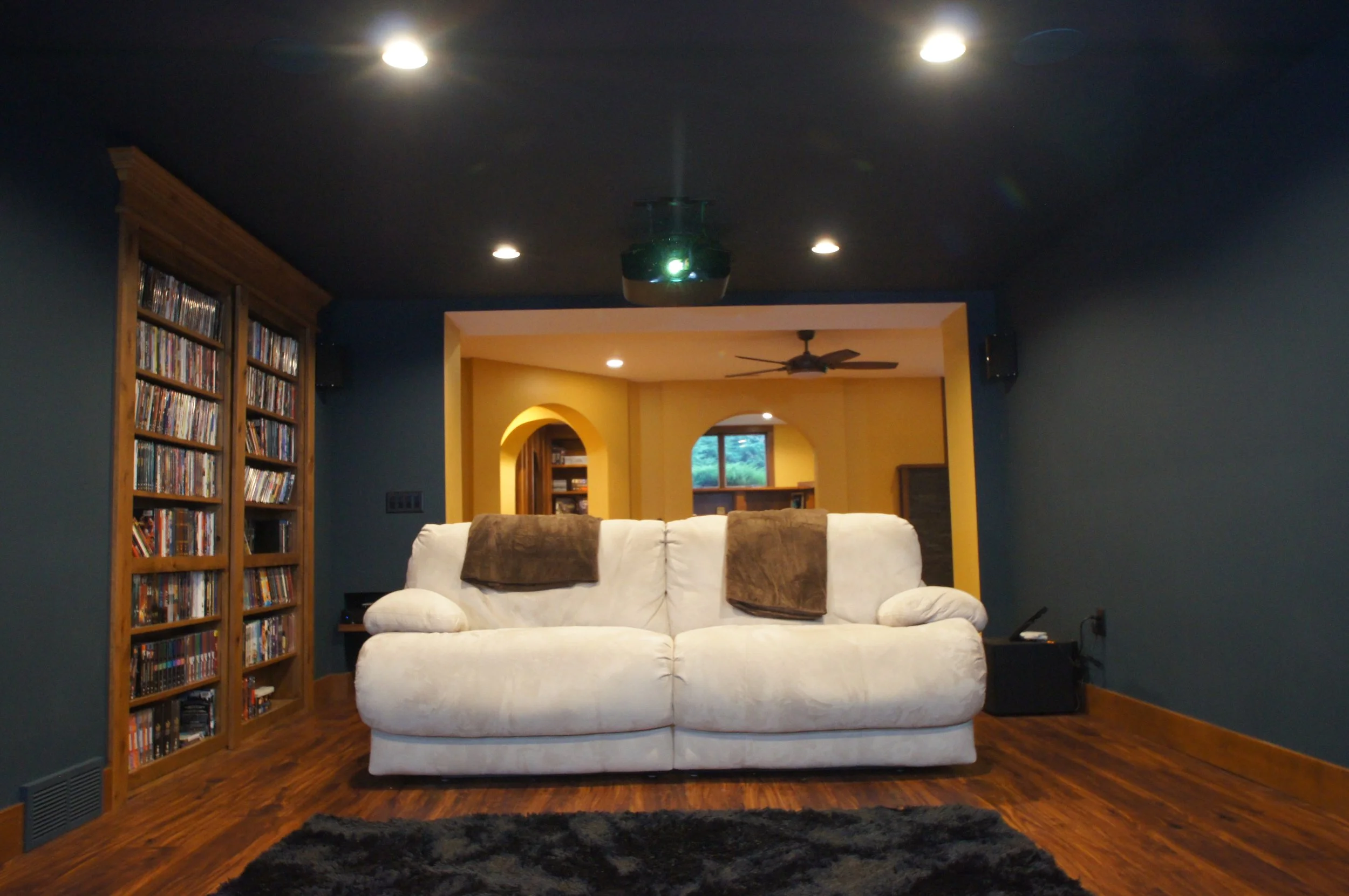 Living room with dark blue walls, a white sofa with brown towels, wooden bookshelves filled with DVDs on the left, hardwood floors, black shag rug, and a doorway in the back leading to a yellow room with a ceiling fan and window.