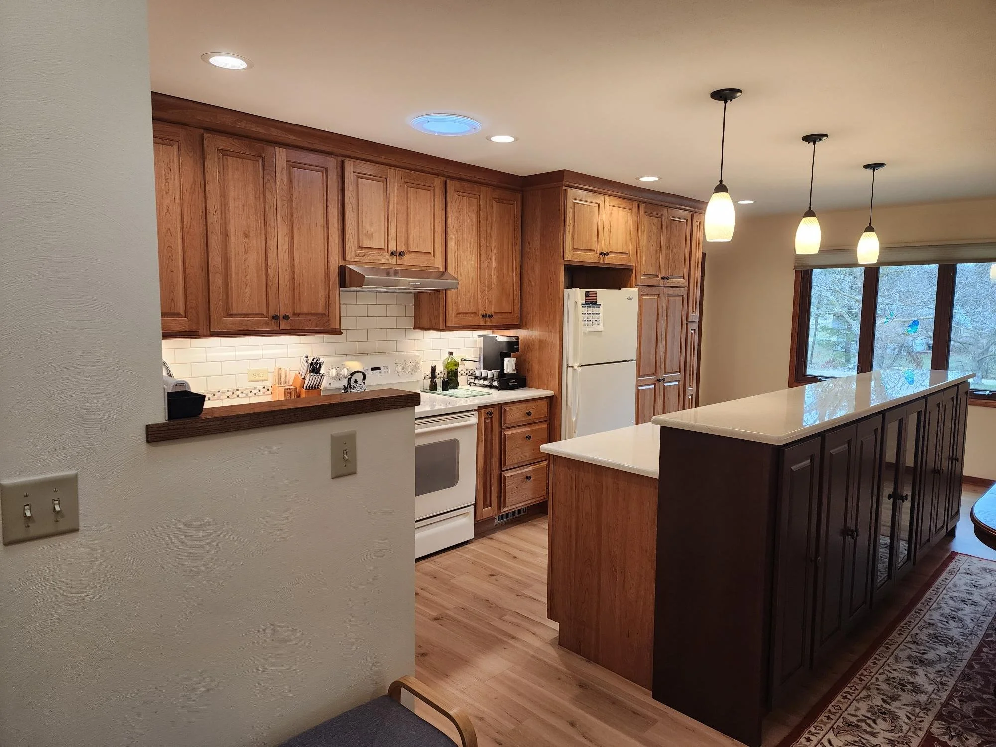 Kitchen with wooden cabinets, white appliances, and a white countertop island. There are pendant lights above the island and a large window providing natural light.