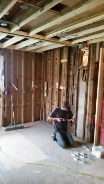 A person kneeling inside a house under construction, working on electrical wiring in the unfinished wooden walls with exposed studs and ceiling.
