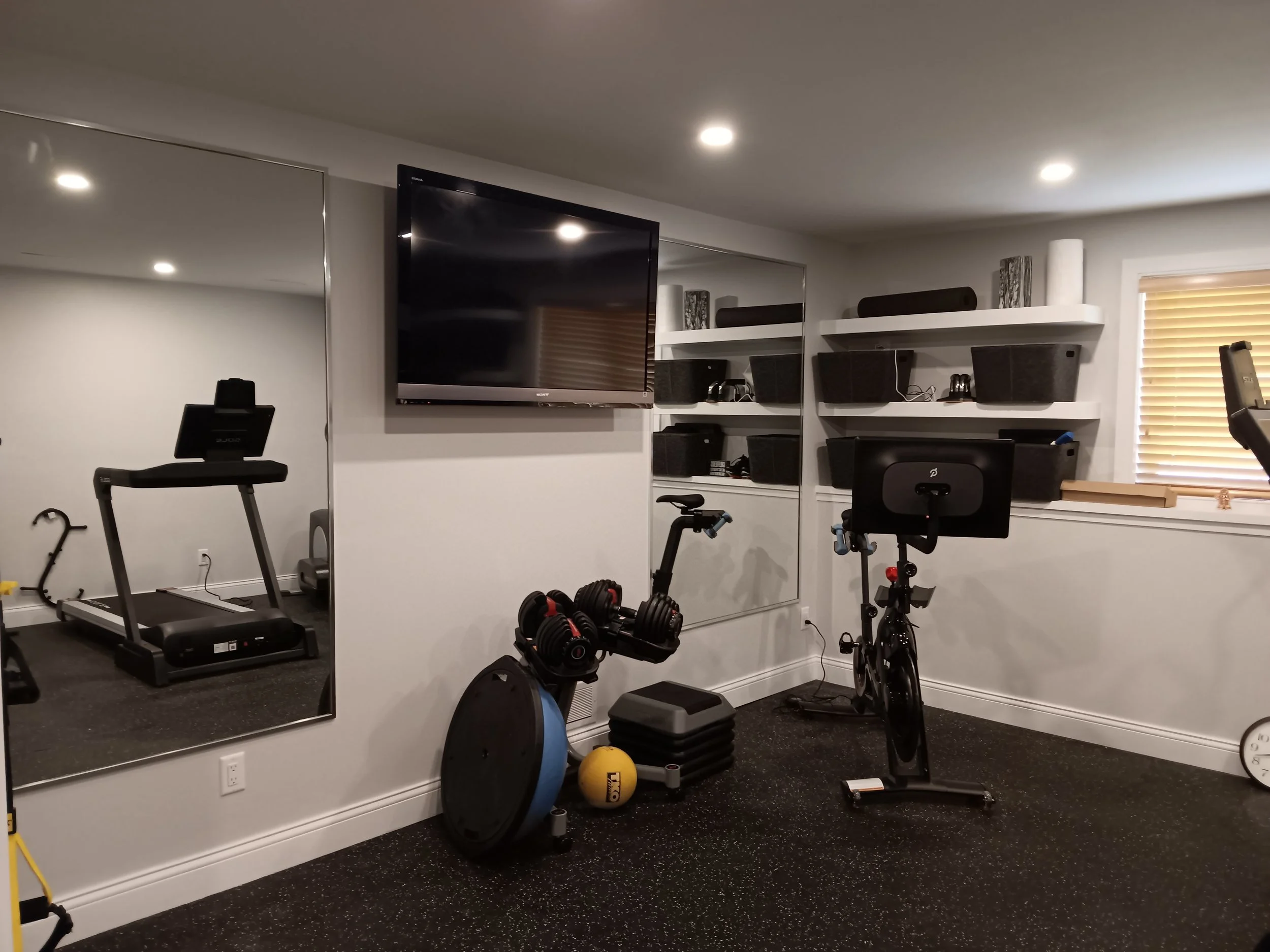 Home gym room with treadmill, exercise bike, dumbbells, and wall-mounted TV with mirrored wall.