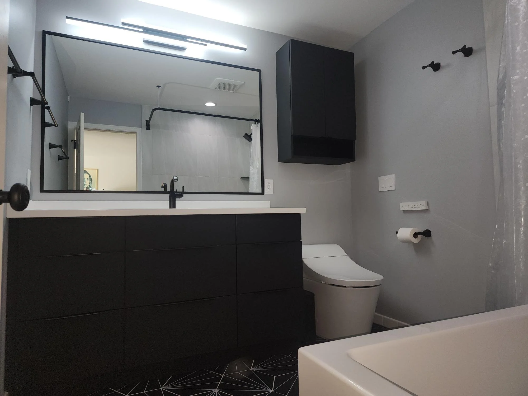 Modern bathroom with black vanity, large mirror, gray walls, black fixtures, black cabinet, white toilet, toilet paper holder, and shower area with curtain. Floor has geometric black and white pattern.