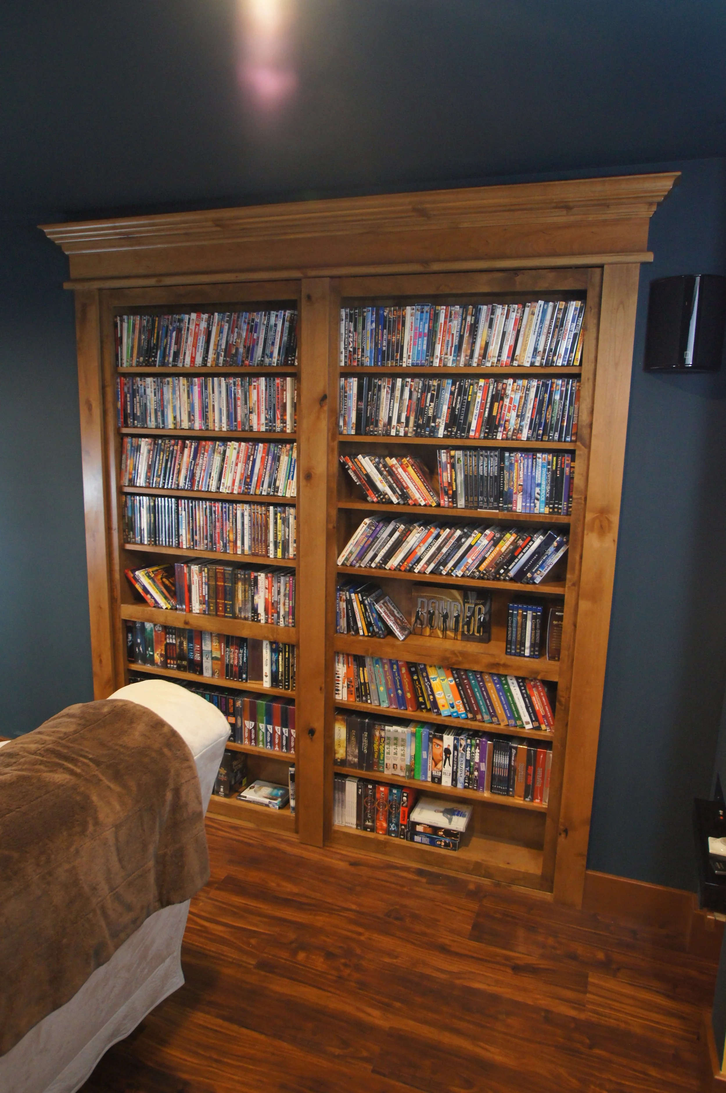 A wooden bookshelf filled with DVDs in a living room with dark blue walls and hardwood flooring.