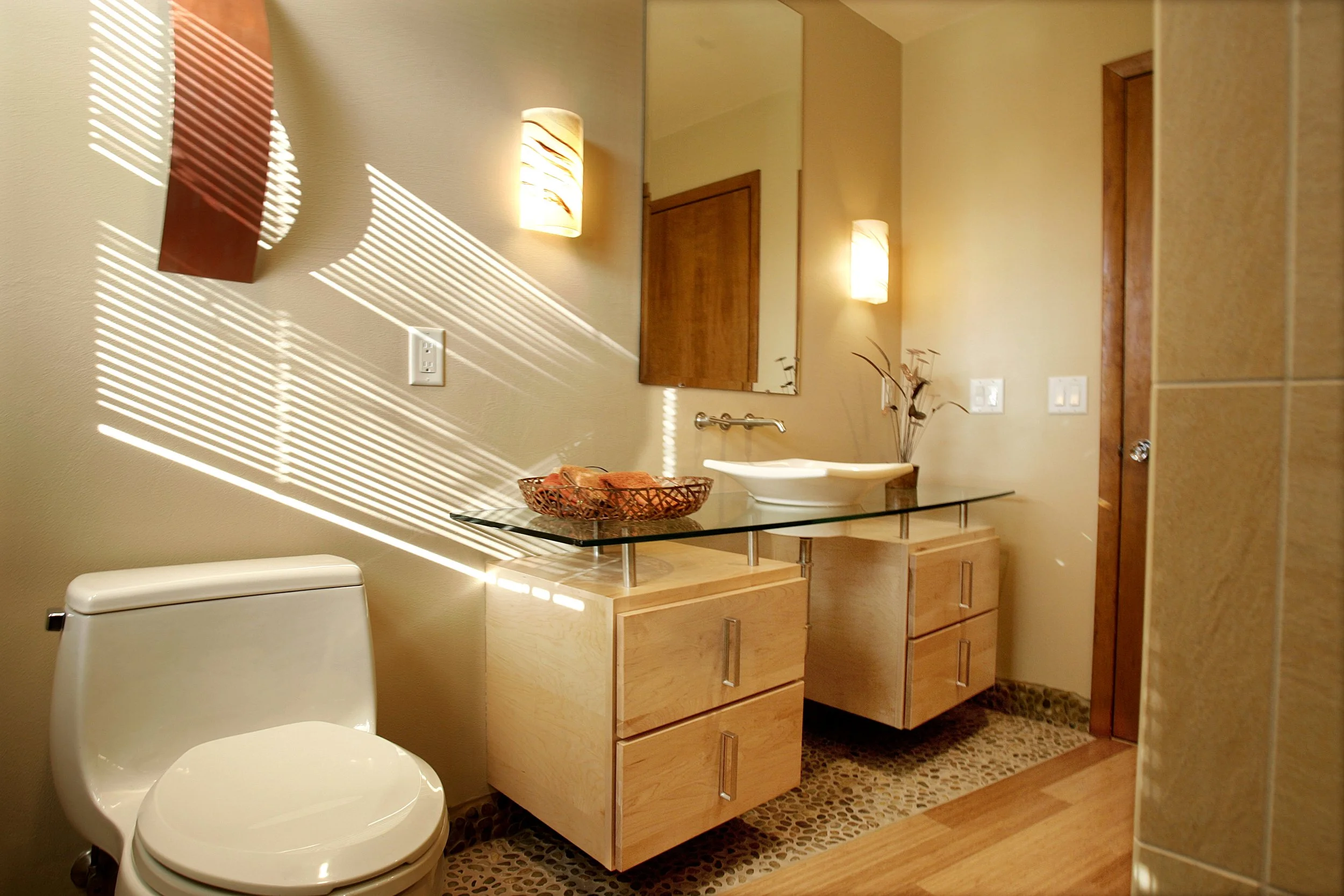 A bathroom with a white toilet, a glass countertop with a vessel sink, a wooden cabinet, wall-mounted lights, a mirror, and decorative items, with sunlight creating striped shadows on the wall.
