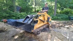 Person operating a yellow bulldozer in a wooded area, pushing dirt or debris.