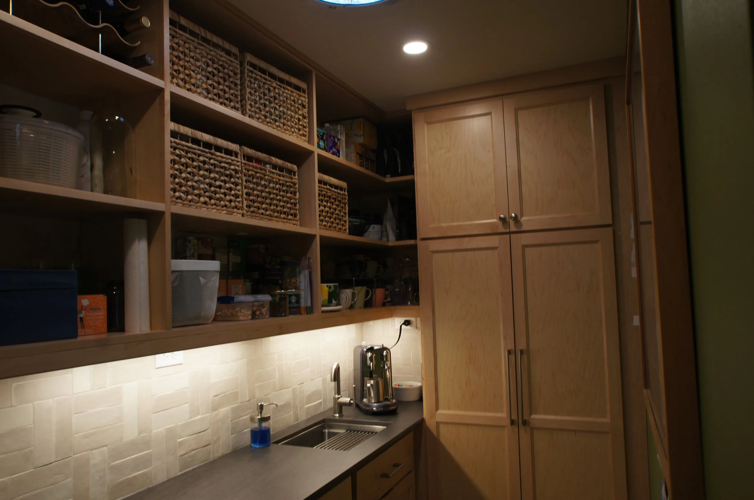 View of a kitchen with wooden cabinets, open shelves with woven baskets, a countertop with a sink, a coffee machine, and a small soap dispenser, illuminated by ceiling lights.