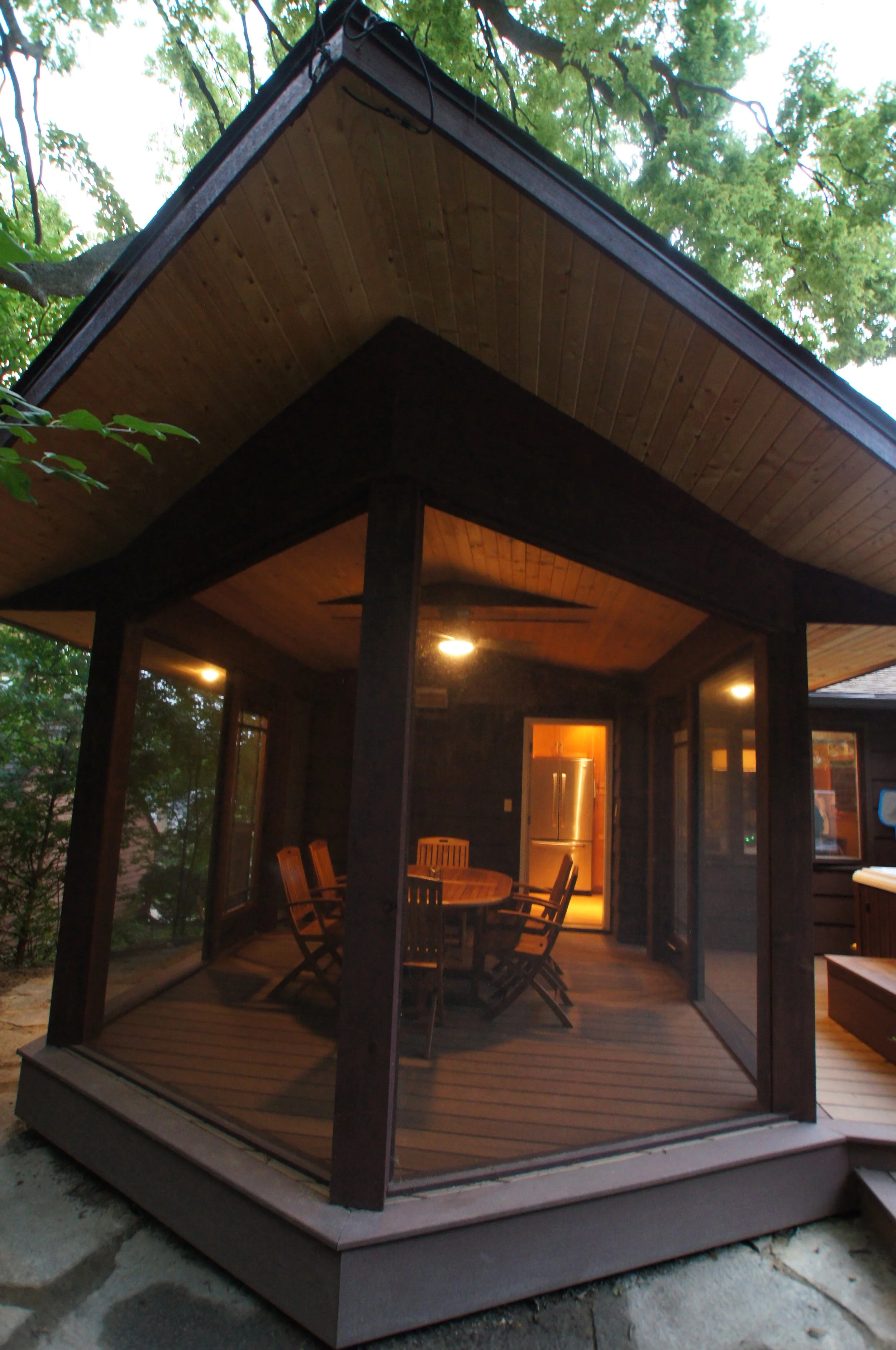 A screened porch with a wooden ceiling, wooden flooring, a round wooden table, and six wooden chairs, with an open door leading to a kitchen with a refrigerator visible inside.