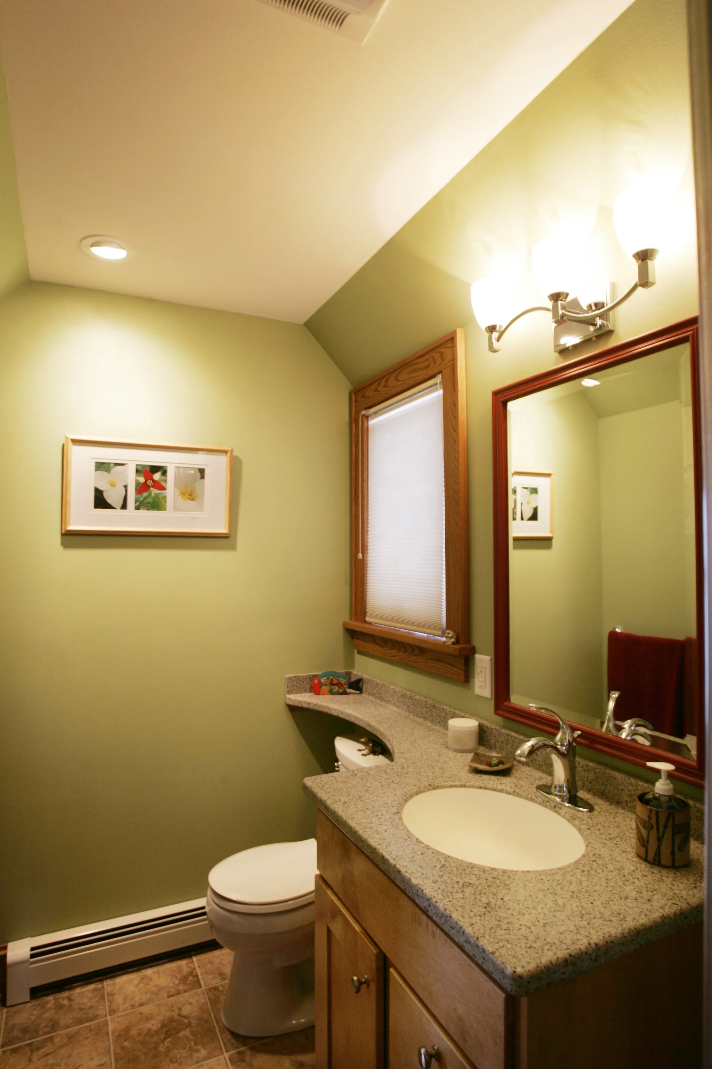 A small bathroom with green walls, a wooden-framed window with a beige shade, a granite sink countertop, a wooden cabinet beneath, a mirror with a wooden frame, and a light fixture with three bulbs above the mirror. There are framed flower pictures o