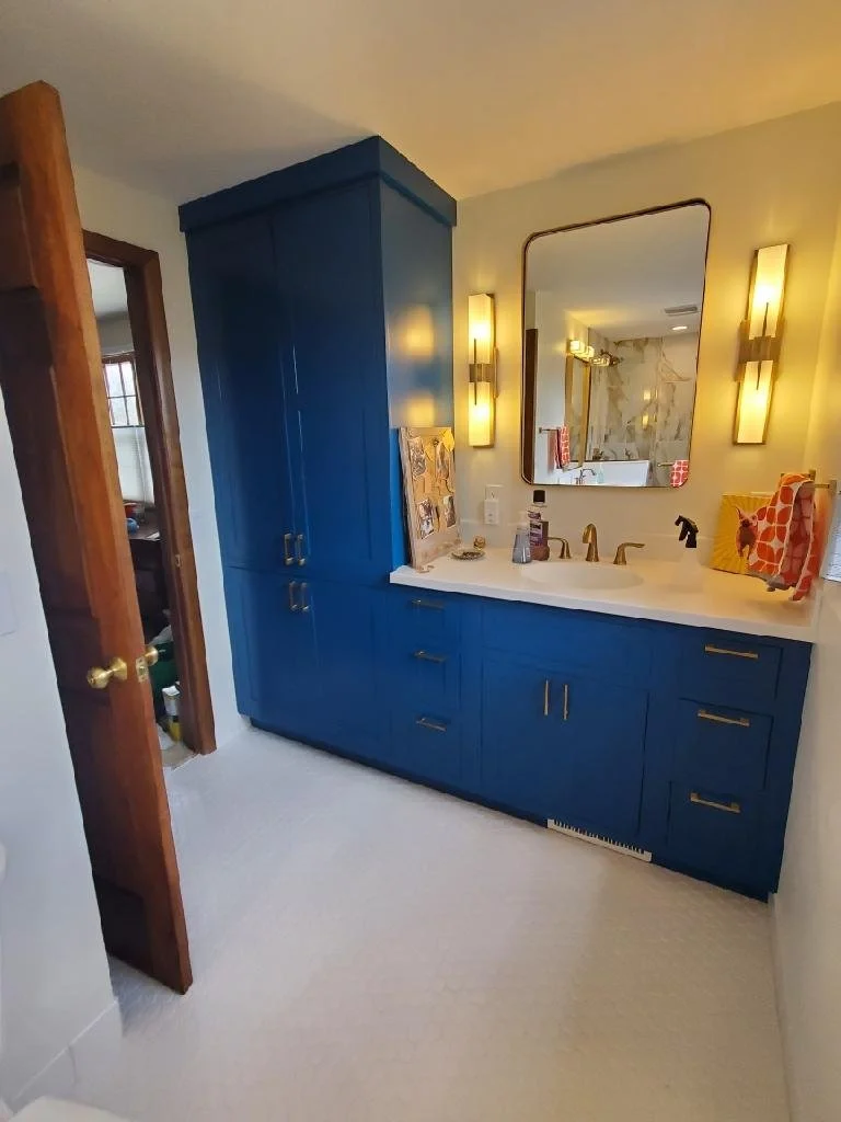 Bathroom vanity with blue cabinets, white countertop, and gold hardware, featuring a large mirror and two vertical wall lights, with a doorway to a walk-in closet