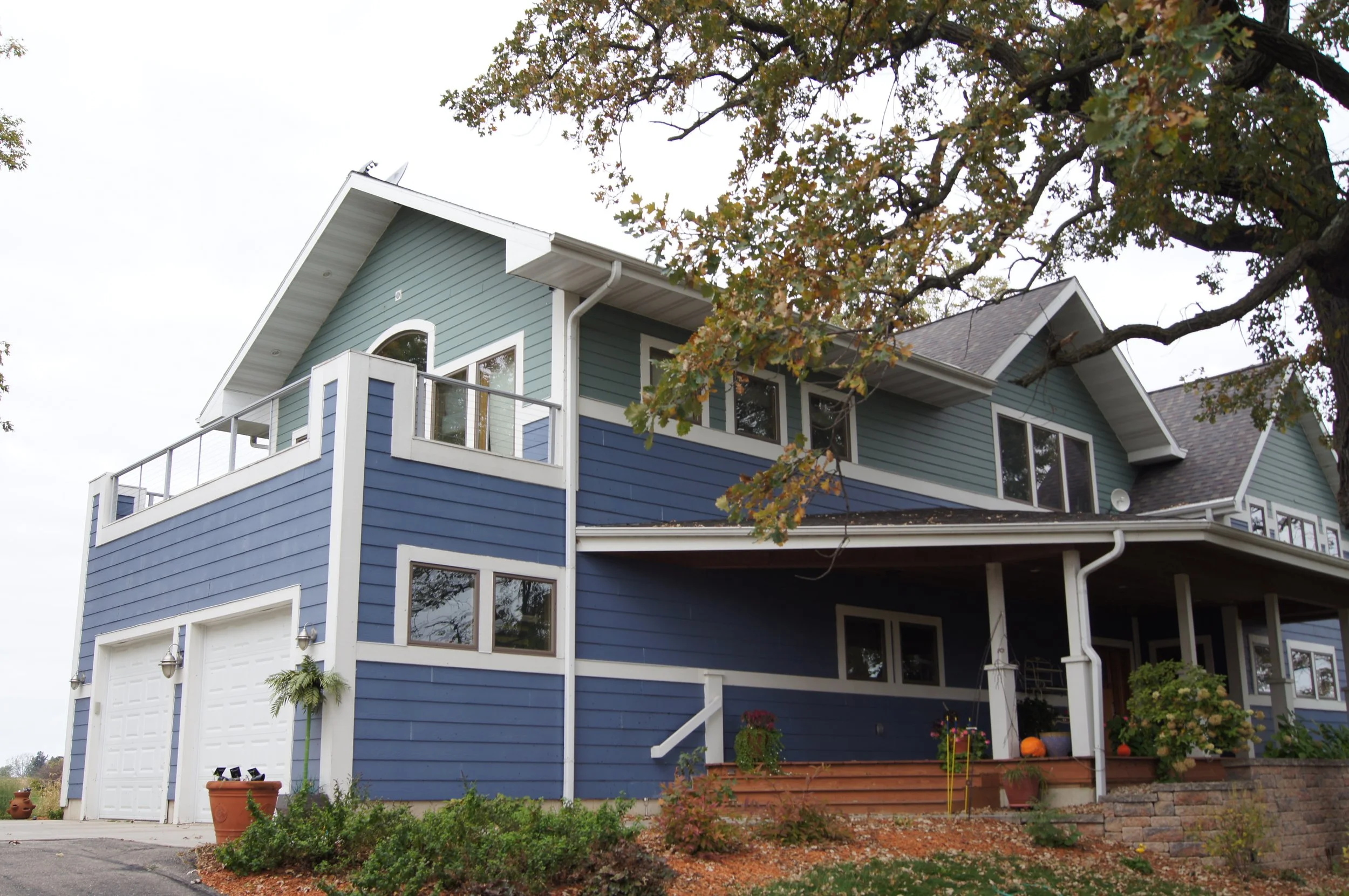 A large blue and green house with white trim, a balcony, and a garage. There are plants and pumpkins near the porch, a tree with fall leaves, and a brick stairway