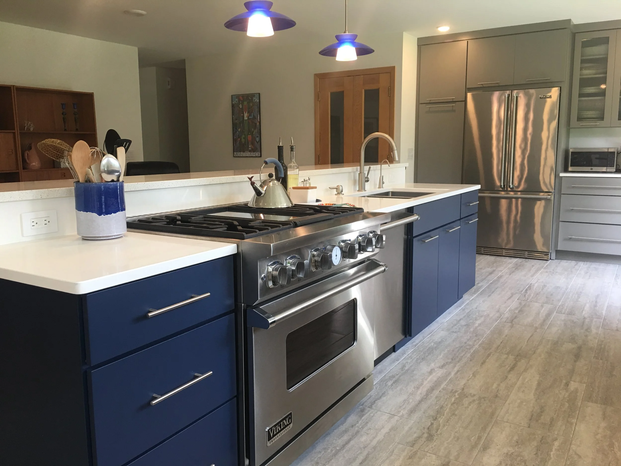 Modern kitchen with blue cabinets, stainless steel appliances, and a white countertop. Items on the counter include a utensil holder with cooking tools, a kettle on the stove, and a small bottle of oil near the sink.