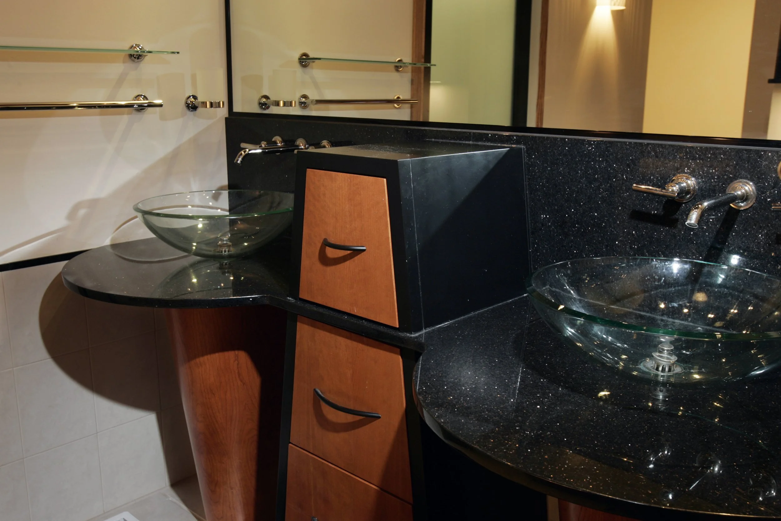 A bathroom vanity with two glass vessel sinks, a black granite countertop, and wooden drawers. A mirror with towel bars is above the sinks, and a wall sconce provides lighting.