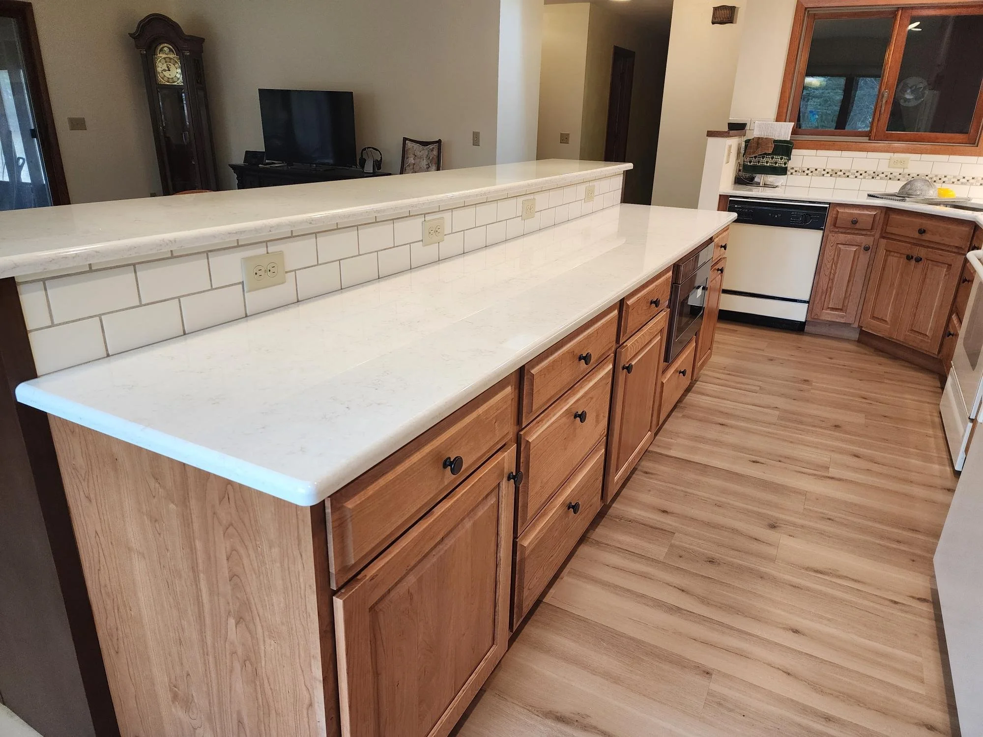 Kitchen with wooden cabinets, white countertops, a white tile backsplash, and hardwood flooring. The view looks over a low wall into a living area with a television and a clock.