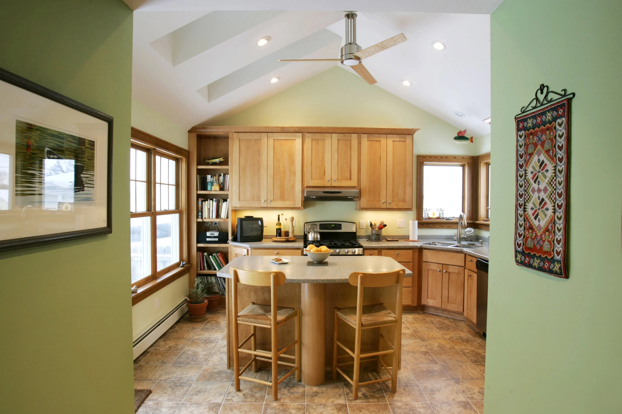 A cozy kitchen with wooden cabinets, a small island with two chairs, and large windows bringing in natural light.