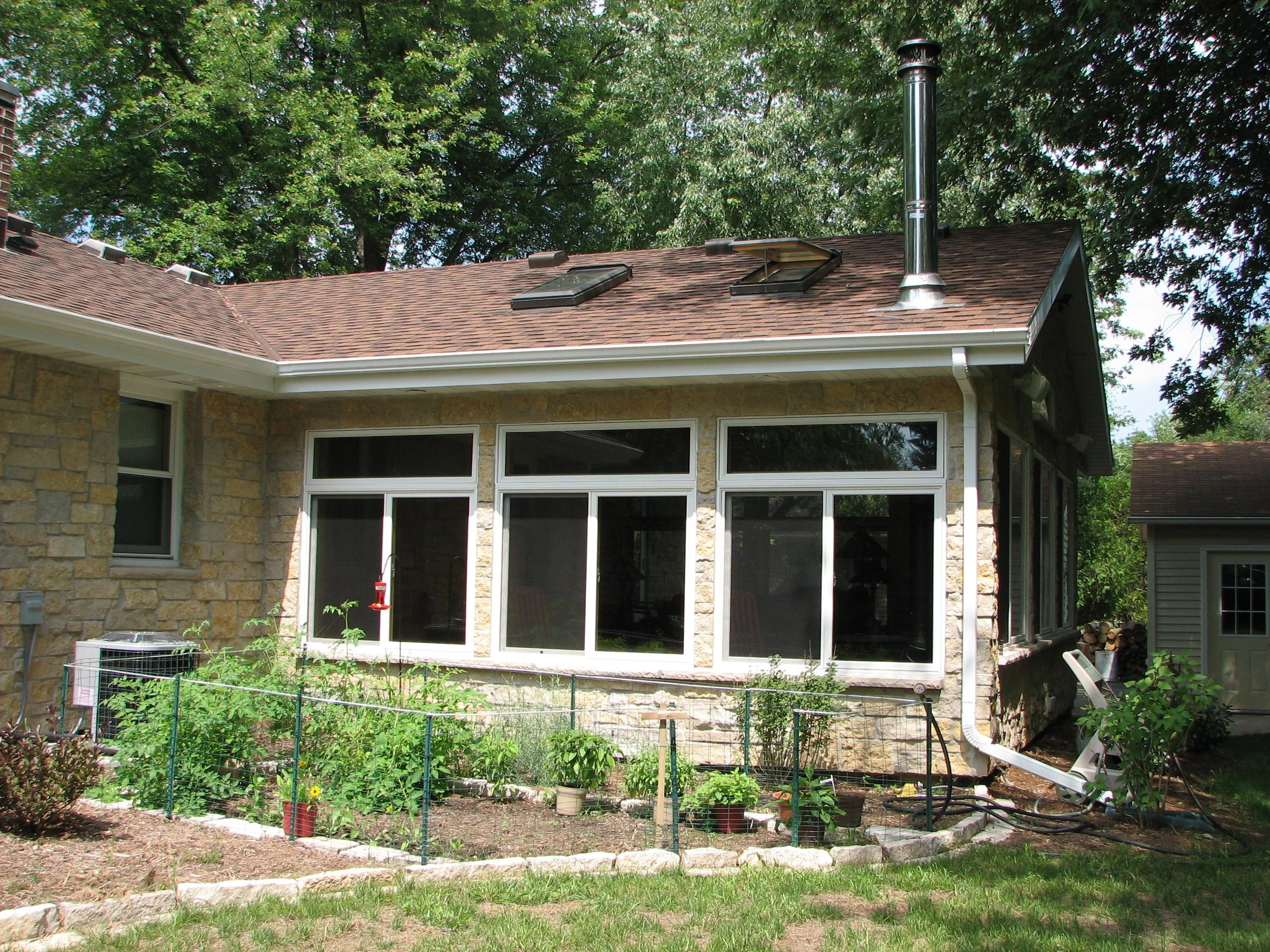 Back of a house with a small garden in front, large windows, stone exterior walls, a shingled roof with skylights and a chimney, surrounded by green trees.