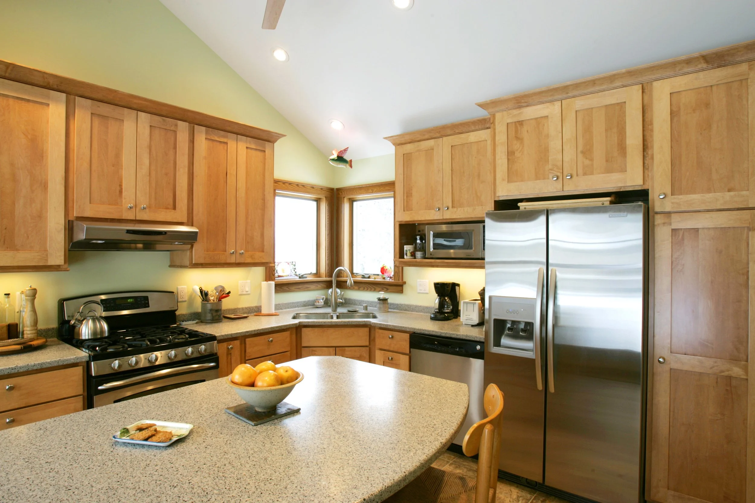 Kitchen with wooden cabinets, stainless steel appliances including a refrigerator, stove, microwave, and dishwasher, a kitchen island with a bowl of apples, and windows above the sink.