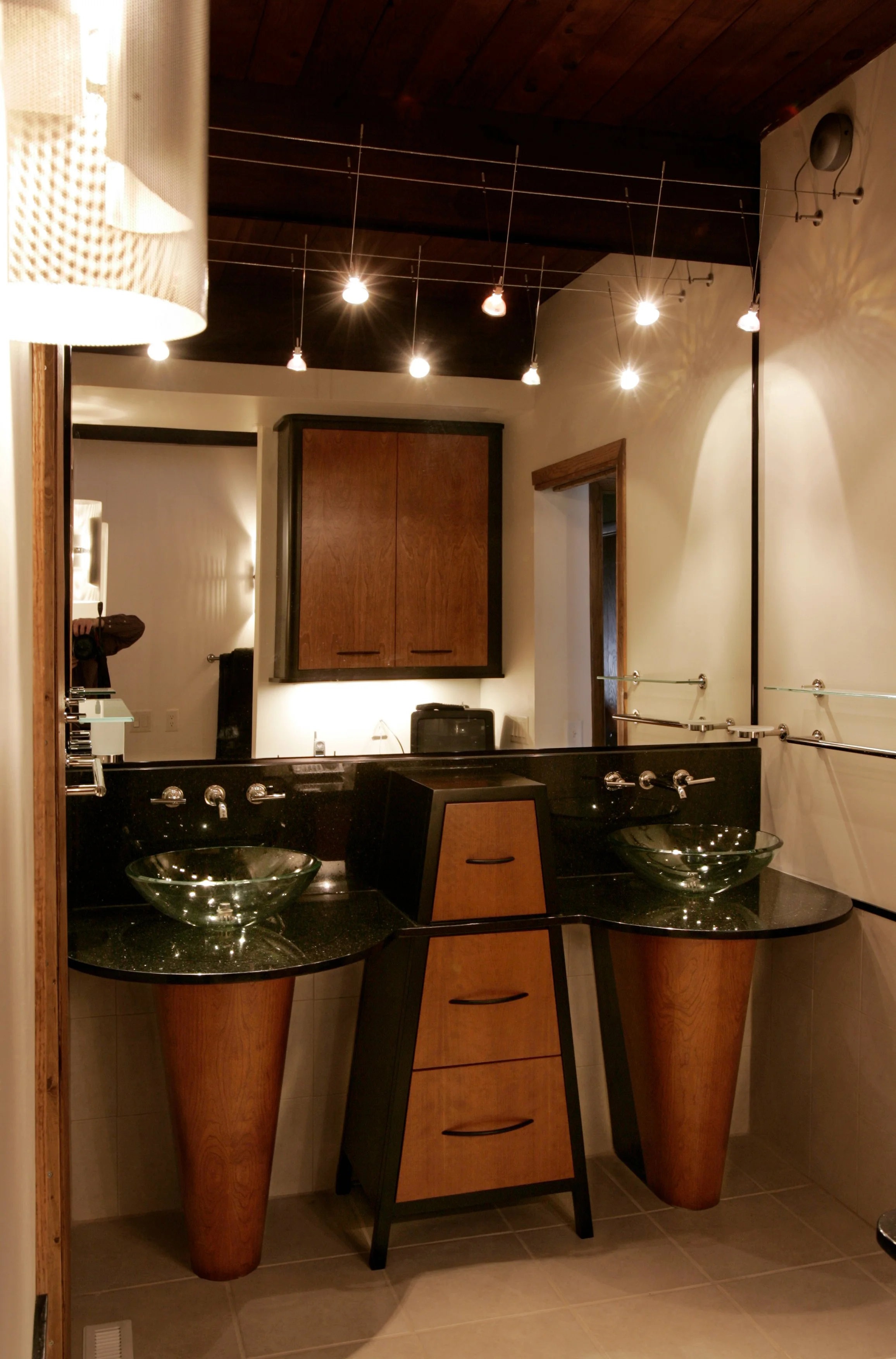 A modern bathroom featuring a black countertop with two glass vessel sinks, a wooden cabinet with three drawers, a large mirror, and ceiling lights.
