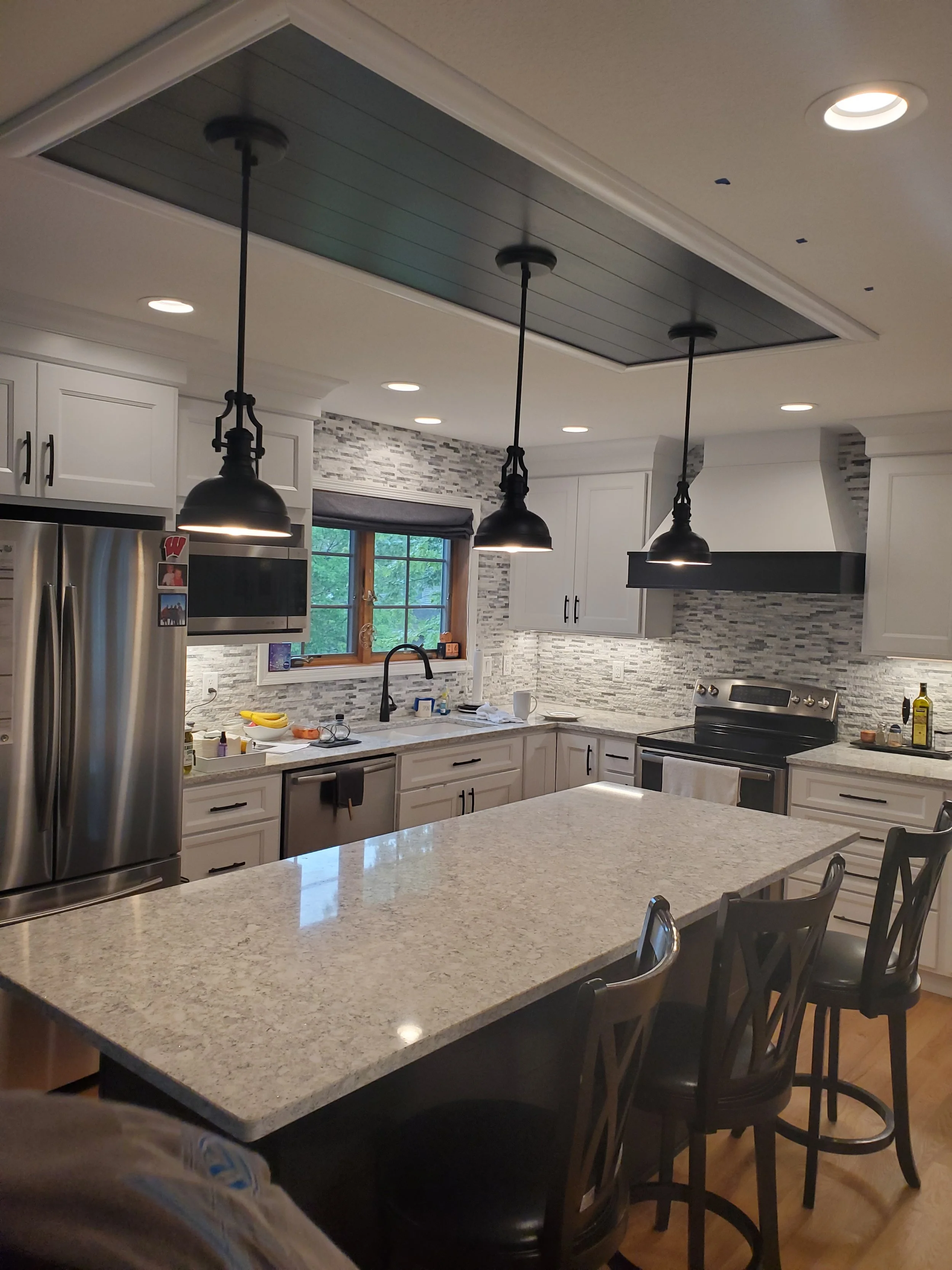 Modern kitchen with white cabinetry, a large granite island, stainless steel appliances, pendant and recessed lighting, a window above the sink, and a backsplash with gray tiles.