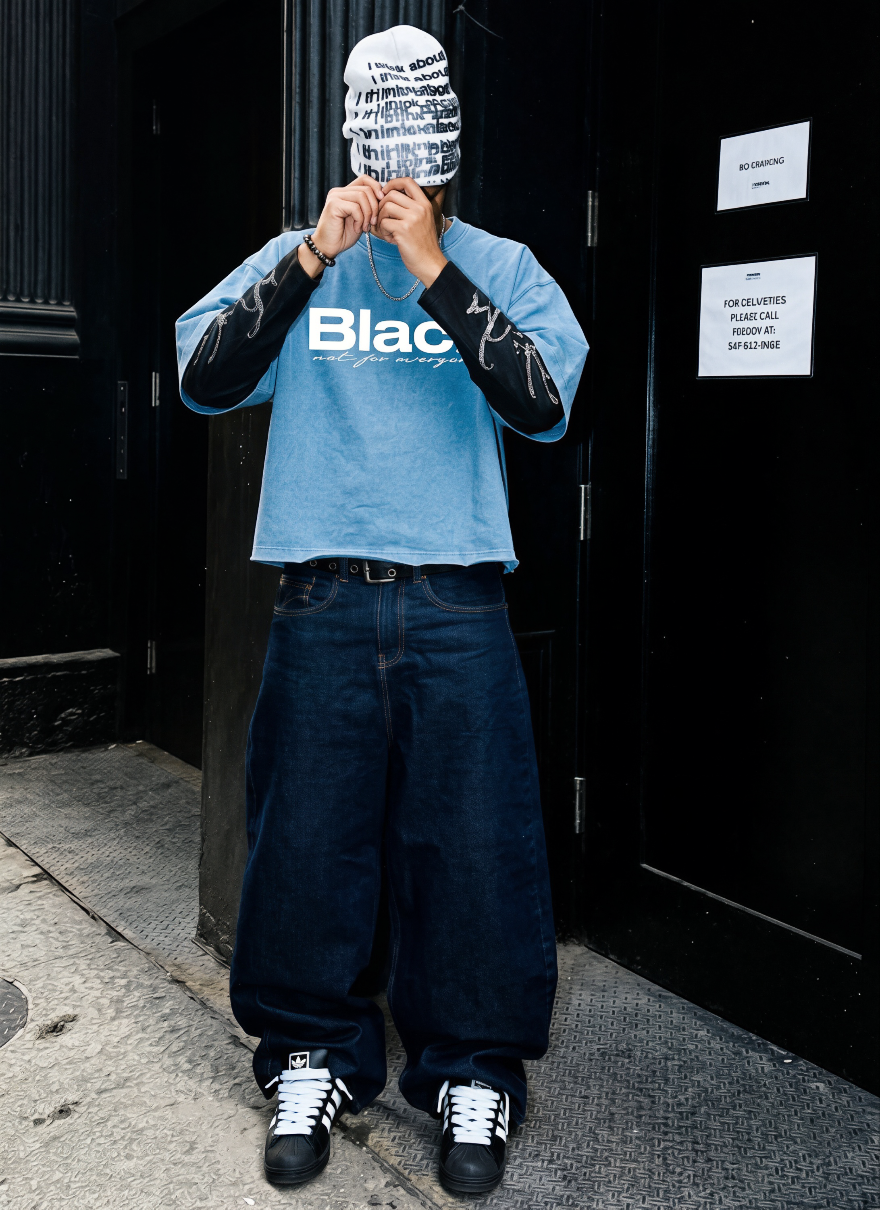 Persona con cabeza cubierta por tela con texto ambiguo, camiseta azul con 'Black' impreso, pantalones anchos y sneakers Adidas, en un entorno urbano oscuro.