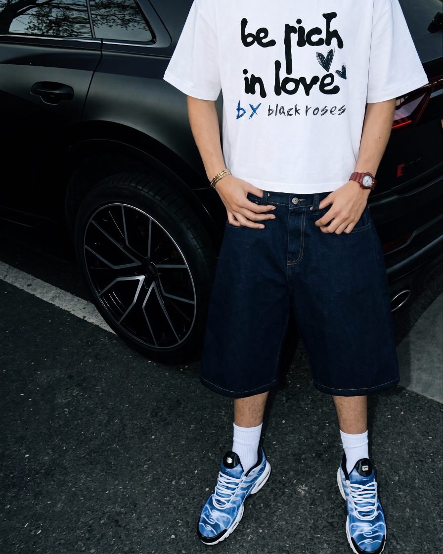 Persona con camiseta blanca con texto 'be rich in love', pantalones cortos, zapatos deportivos y reloj, de pie junto a un coche negro en aparcamiento.