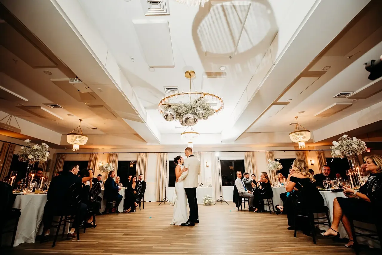 A bride and groom sharing their first dance at their wedding reception in a decorated banquet hall. Guests are seated at round tables on either side, with floral centerpieces and candles.