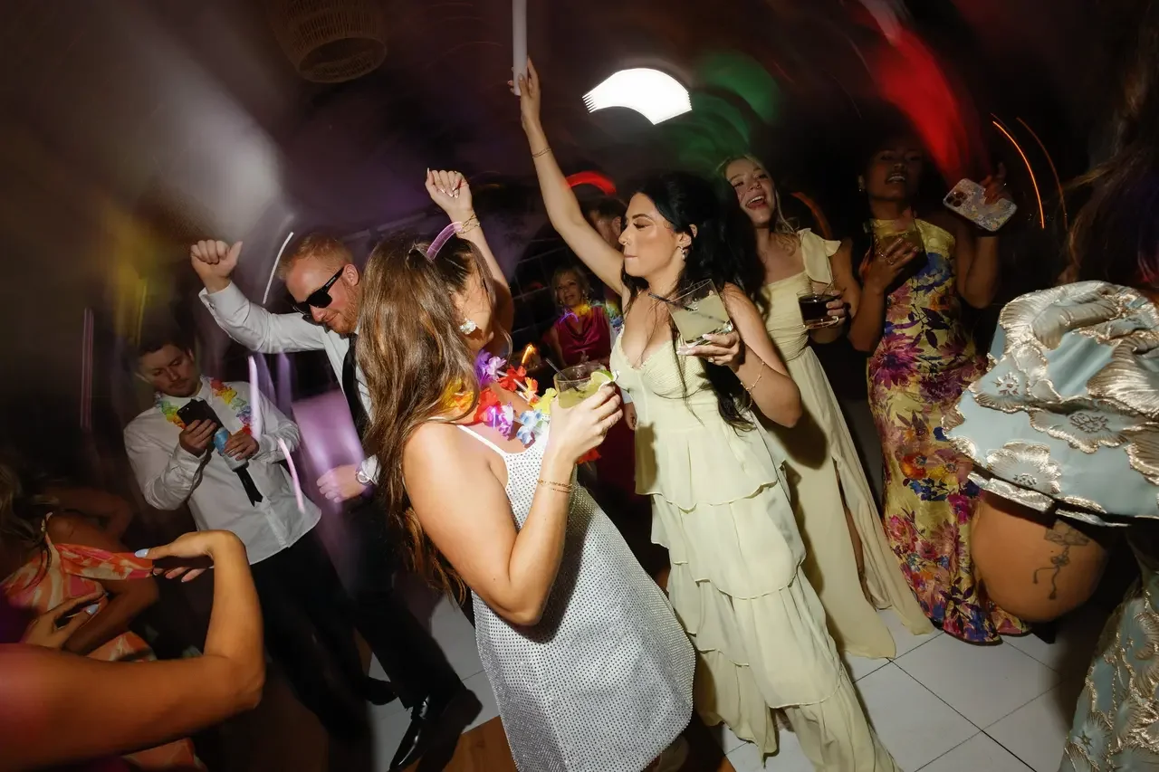 People dancing, drinking, and celebrating at a lively party or celebration, with some wearing leis and colorful dresses.