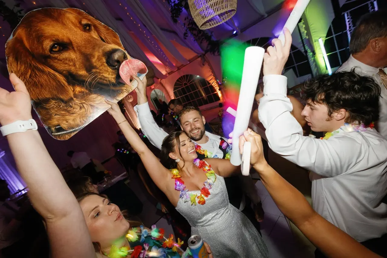 Group of people celebrating at a party, with one person holding a dog face cutout and another holding colorful glow sticks, dancing and smiling under colorful lights.