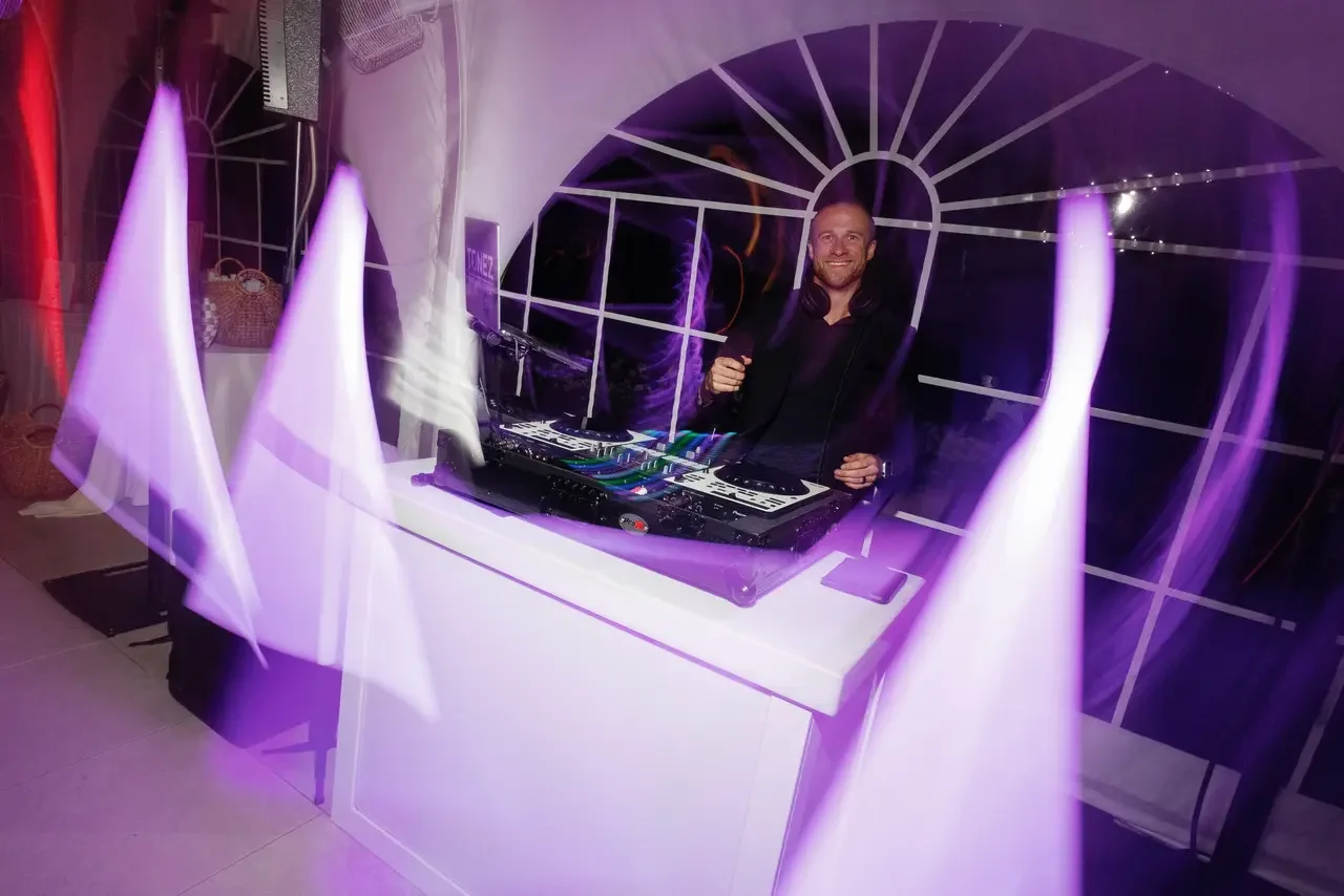 A DJ with headphones around his neck at a DJ booth, smiling, in a party setting with purple lighting.
