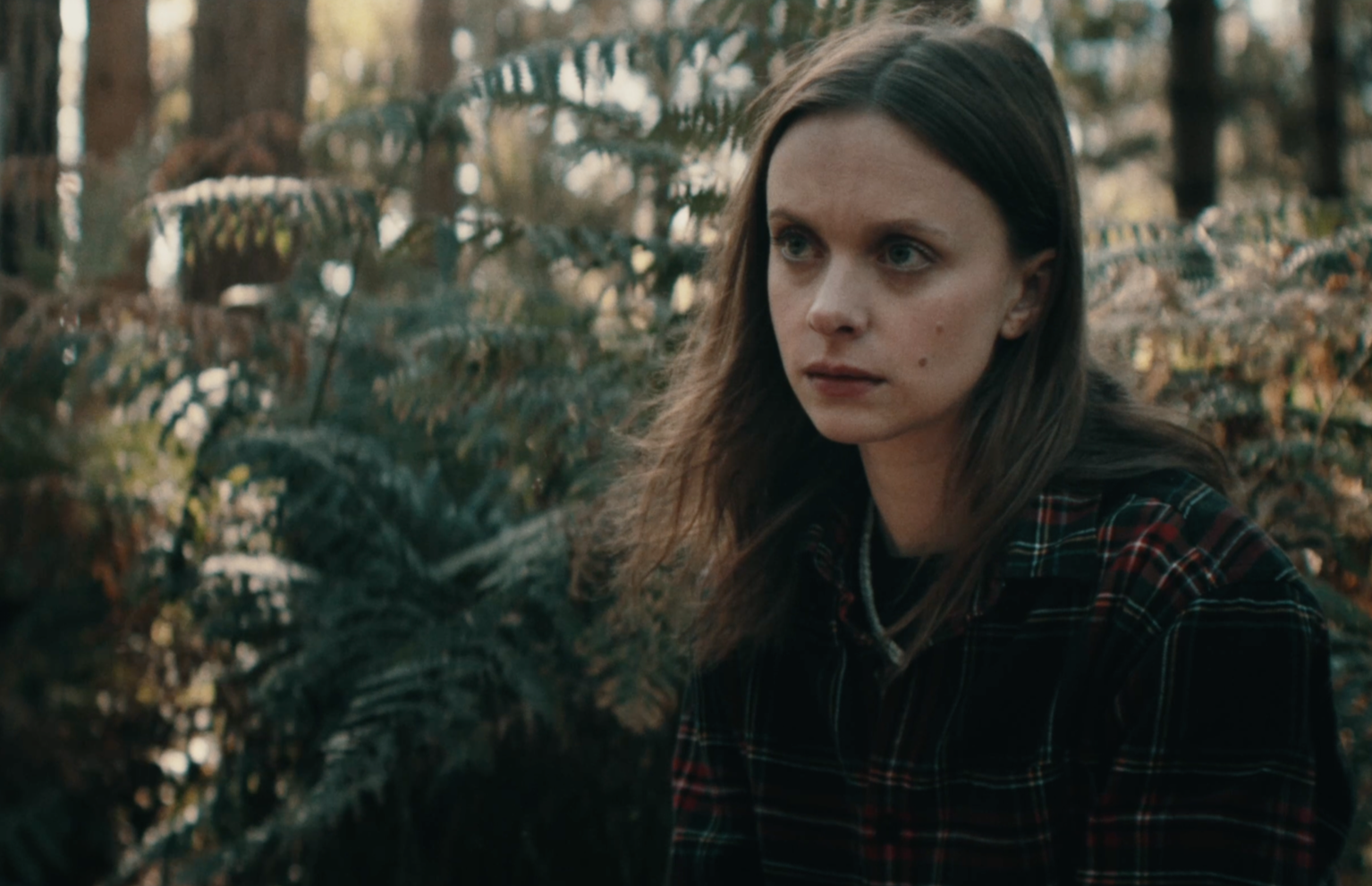 A woman with long brown hair and wearing a black and red plaid shirt, sitting outdoors in a wooded area with ferns and trees in the background.