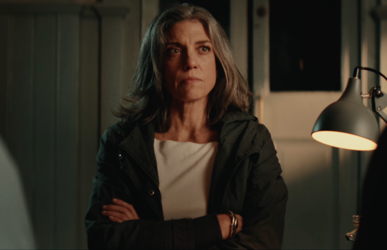 A middle-aged woman with shoulder-length gray hair, wearing a black jacket and a white shirt, standing with her arms crossed in a room with wooden paneling and a small desk lamp.