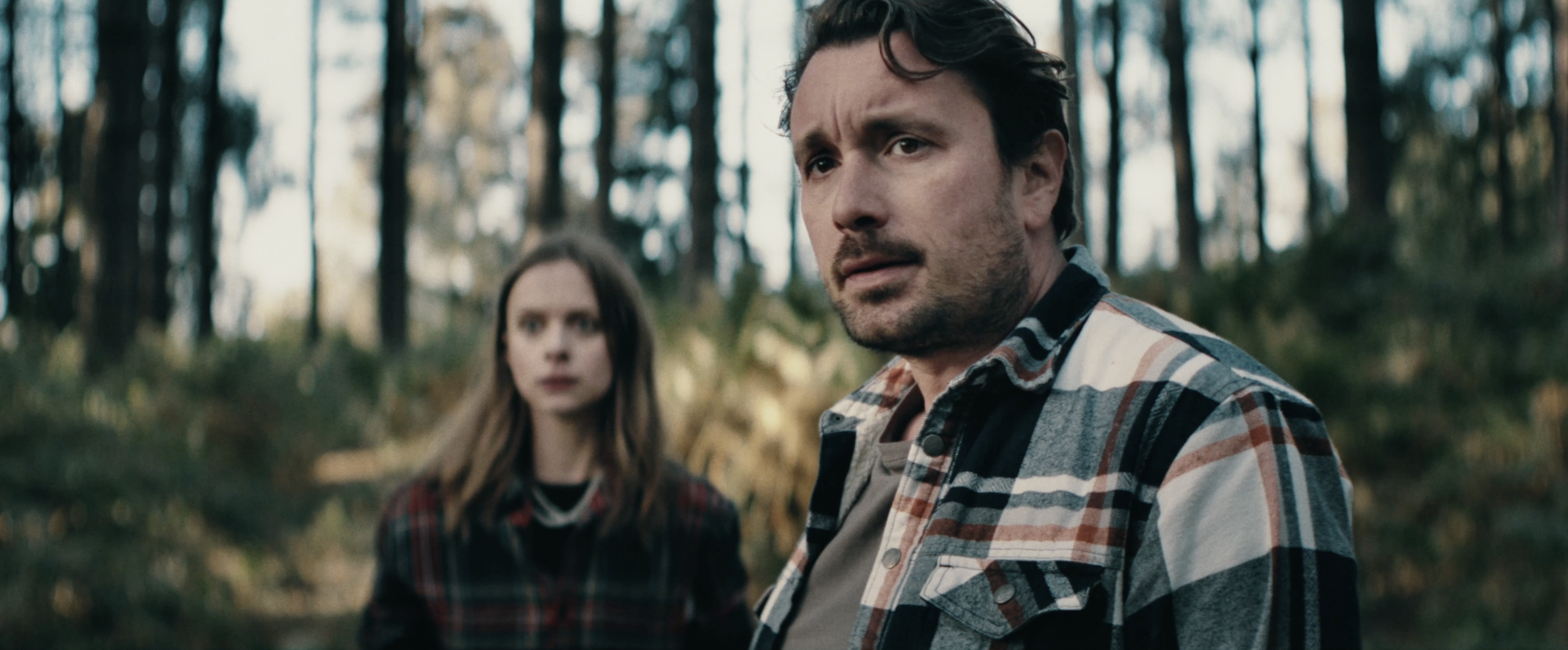 A man with dark hair and a woman with light brown hair in a forest setting, both looking serious or concerned. The man is in the foreground, wearing a plaid shirt, and the woman is in the background, also wearing a plaid shirt.