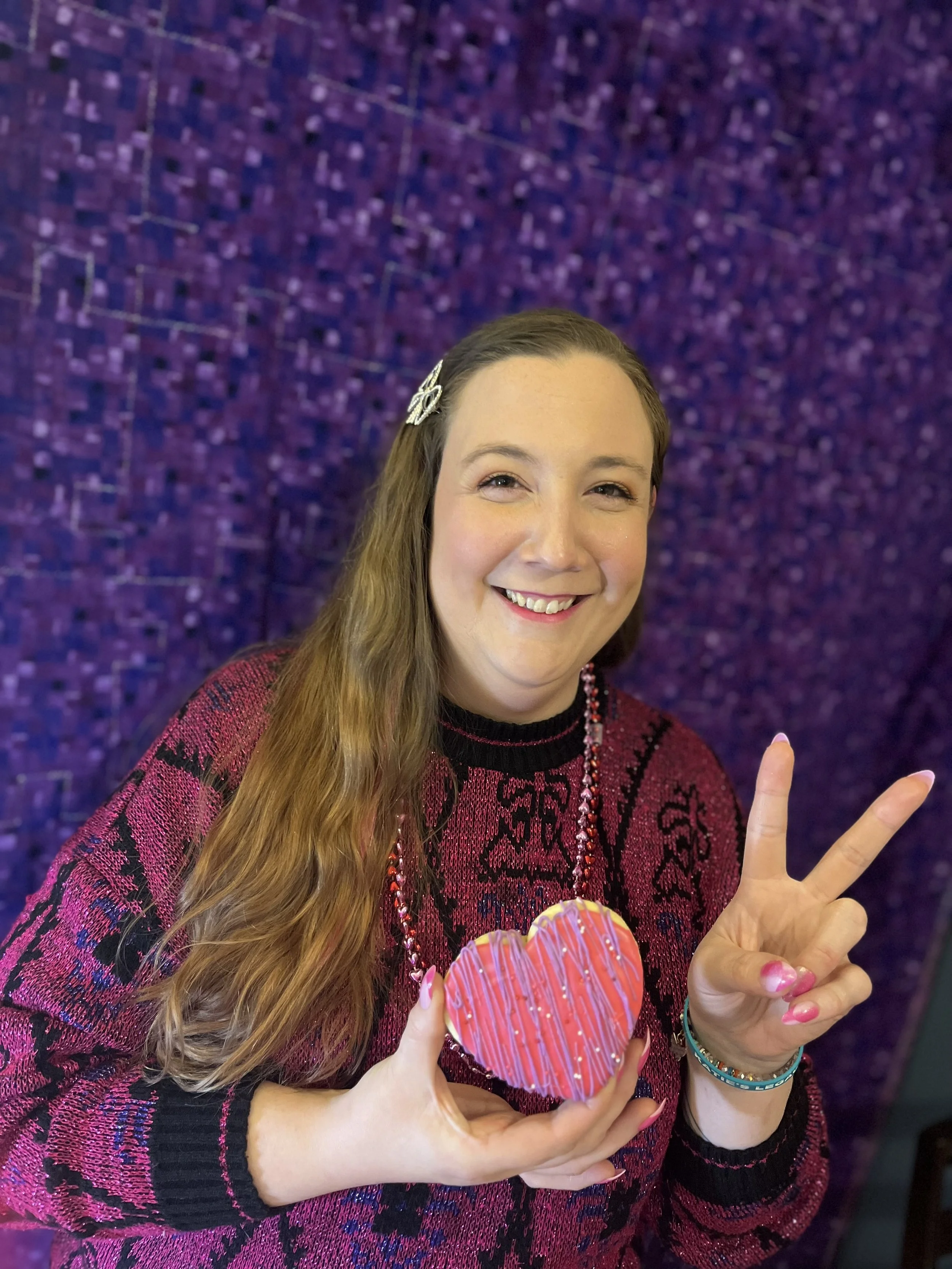A woman with long wavy hair, wearing a pink and black sweater and jewelry, smiling, holding a decorated heart-shaped cookie and making a peace sign with her other hand, standing in front of a purple wall.