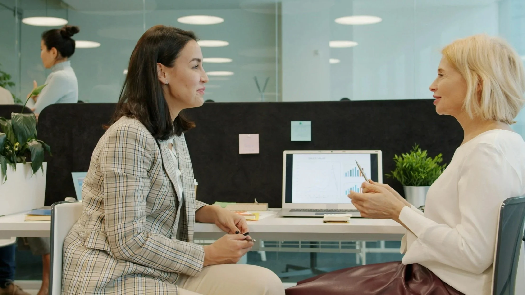 Two women sit across from each other at a desk in an office, engaging in a conversation. One woman has dark hair and is wearing a plaid blazer, while the other woman has blonde hair and is dressed in a white top. They are smiling and holding pens, with a laptop displaying sales data and charts on the desk between them. A woman in a white blouse with a bun is blurred in the background, working at another desk.