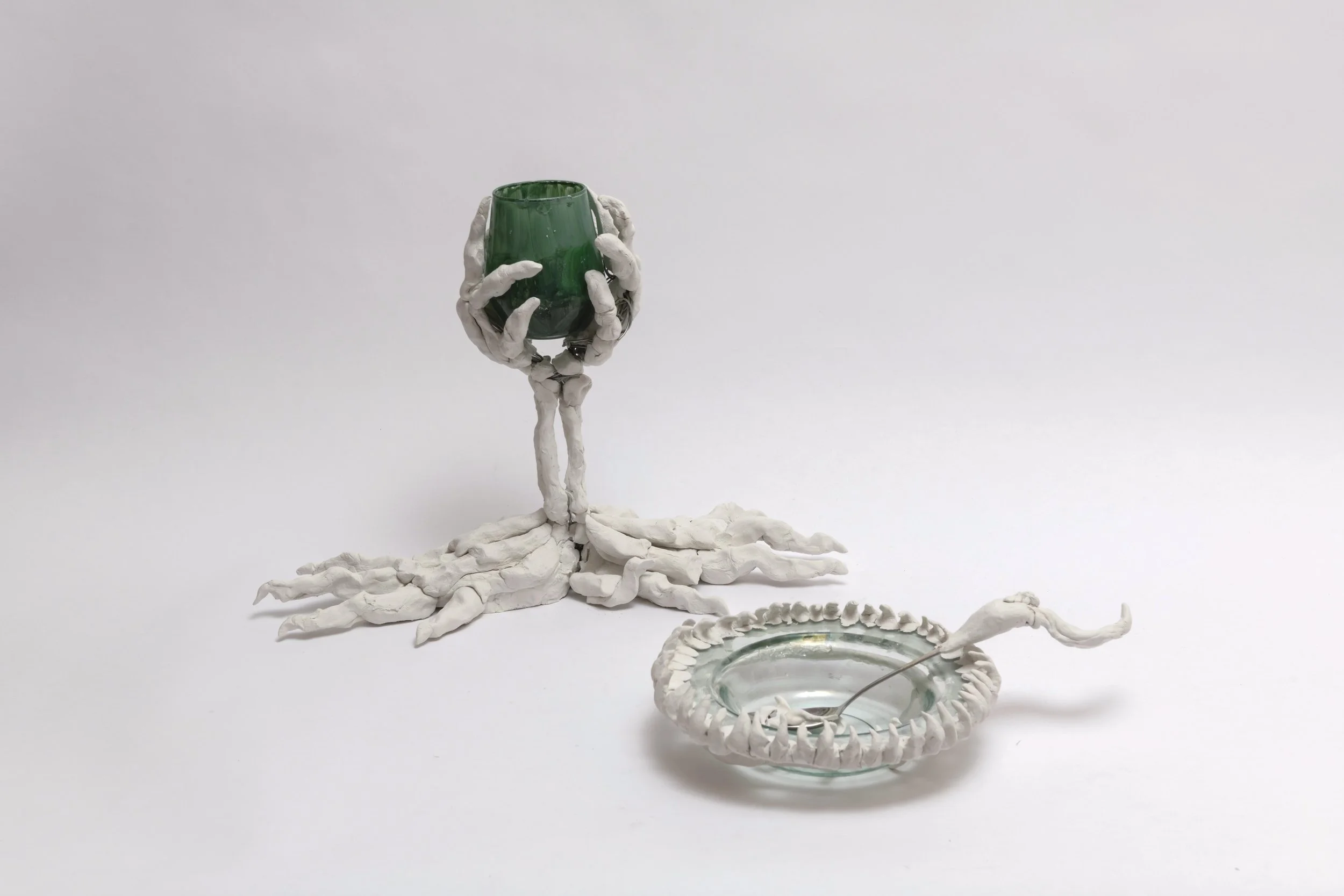 Artistic sculpture of human hands made from a white material, one hand holding a green glass, and another hand holding a spoon in a glass bowl with a white rim, all set against a plain white background.