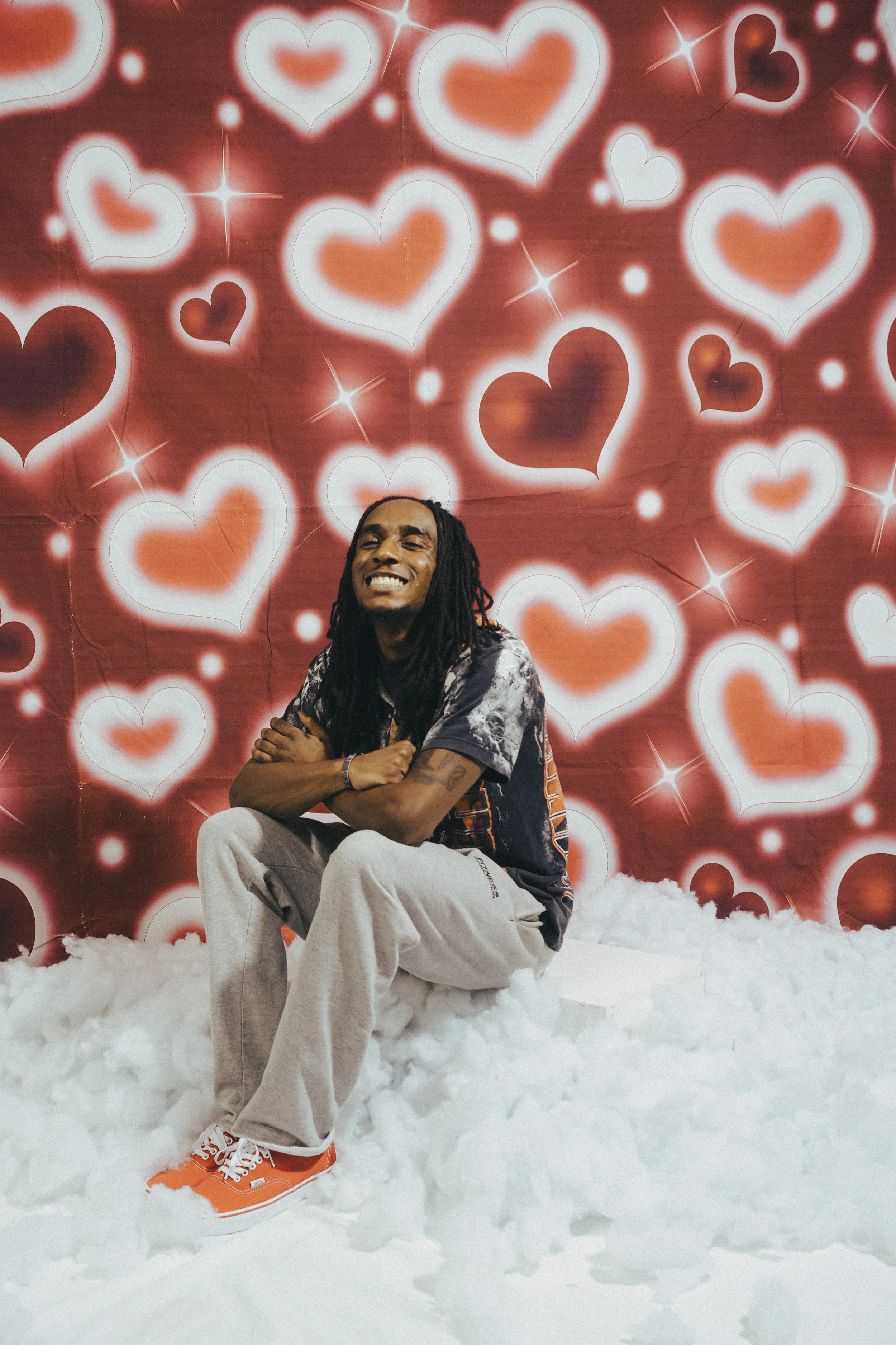 A young man with dreadlocks smiling and sitting on a white platform surrounded by fluffy white material, with a background of red and white glowing hearts and starbursts.