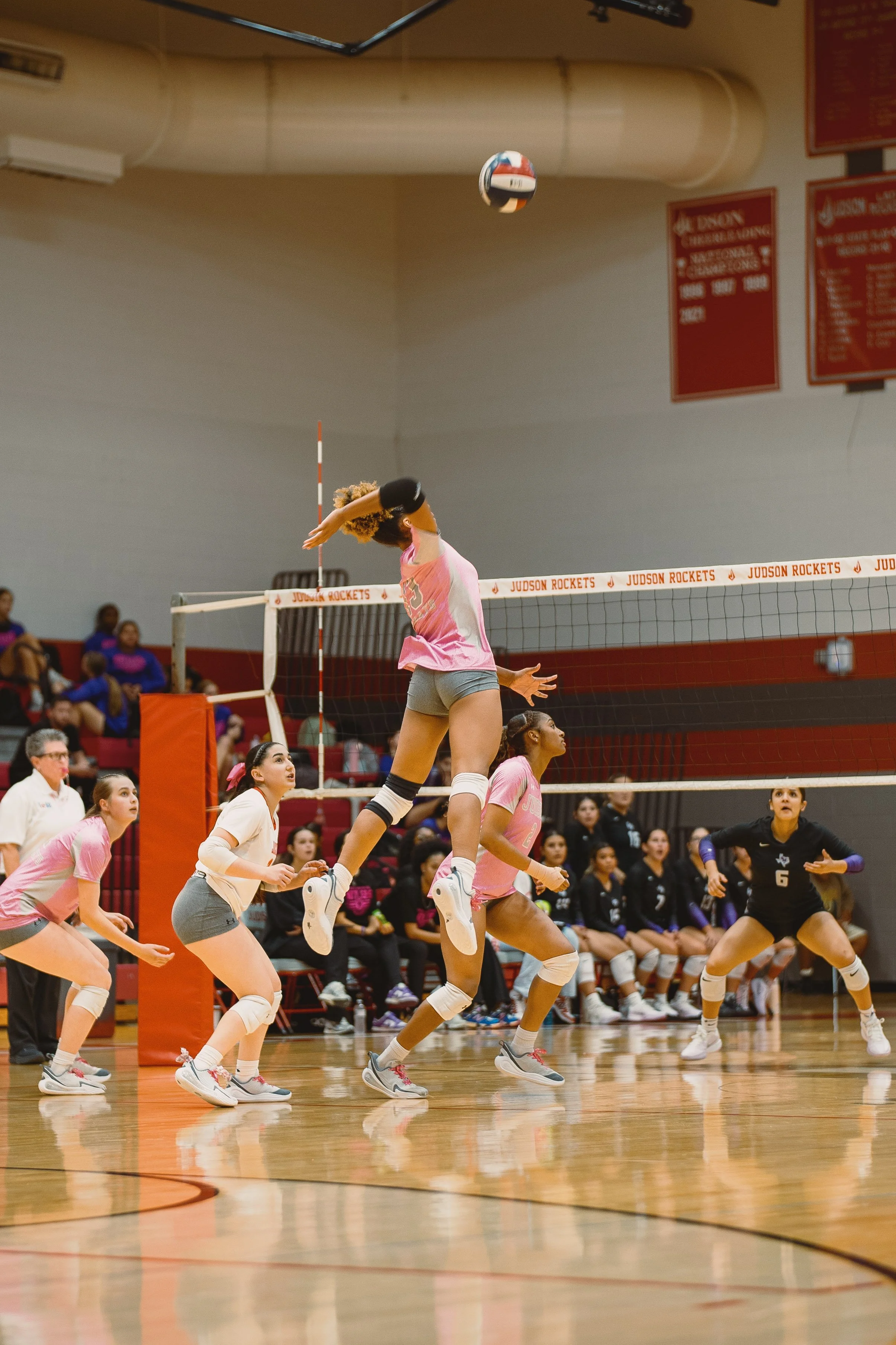 A women's volleyball game where a player in pink jumps to hit the ball over the net, with teammates and opponents watching.