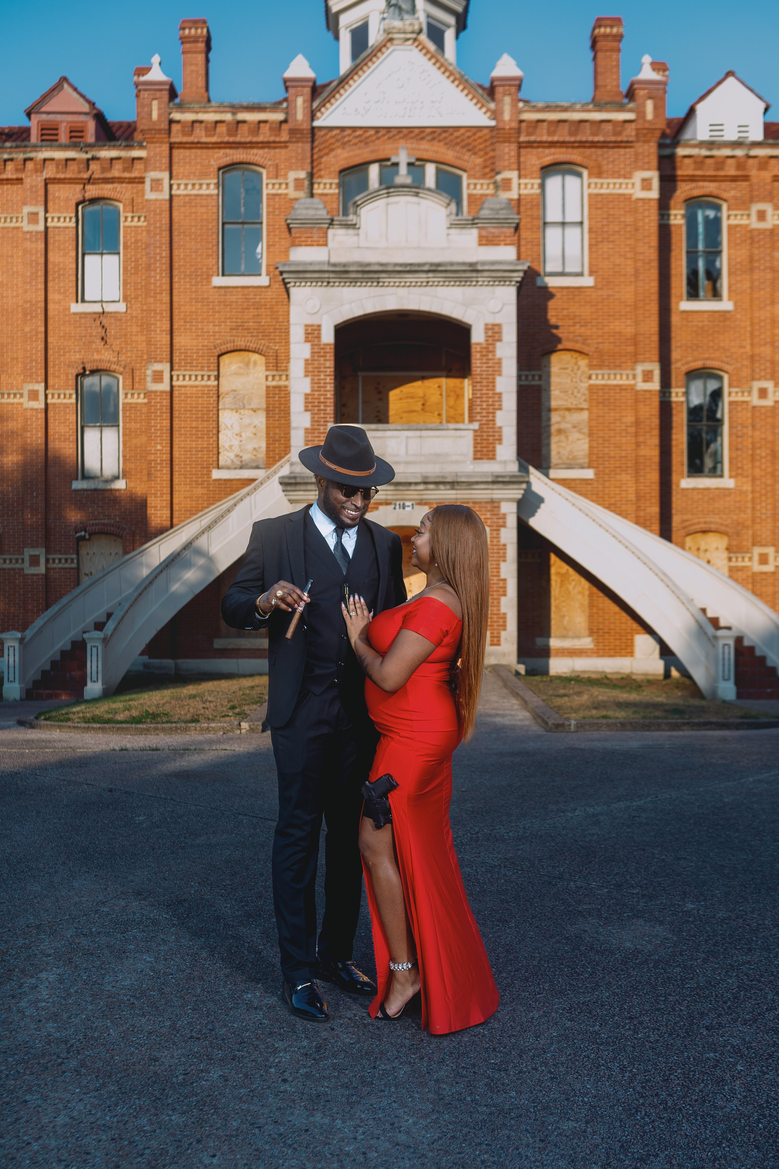 A man and woman standing together on a street in front of a large, red brick building with stairs and archways. The man is wearing a suit, tie, and fedora, while the woman is in a long, red dress with black high heels and a garter belt showing. They 