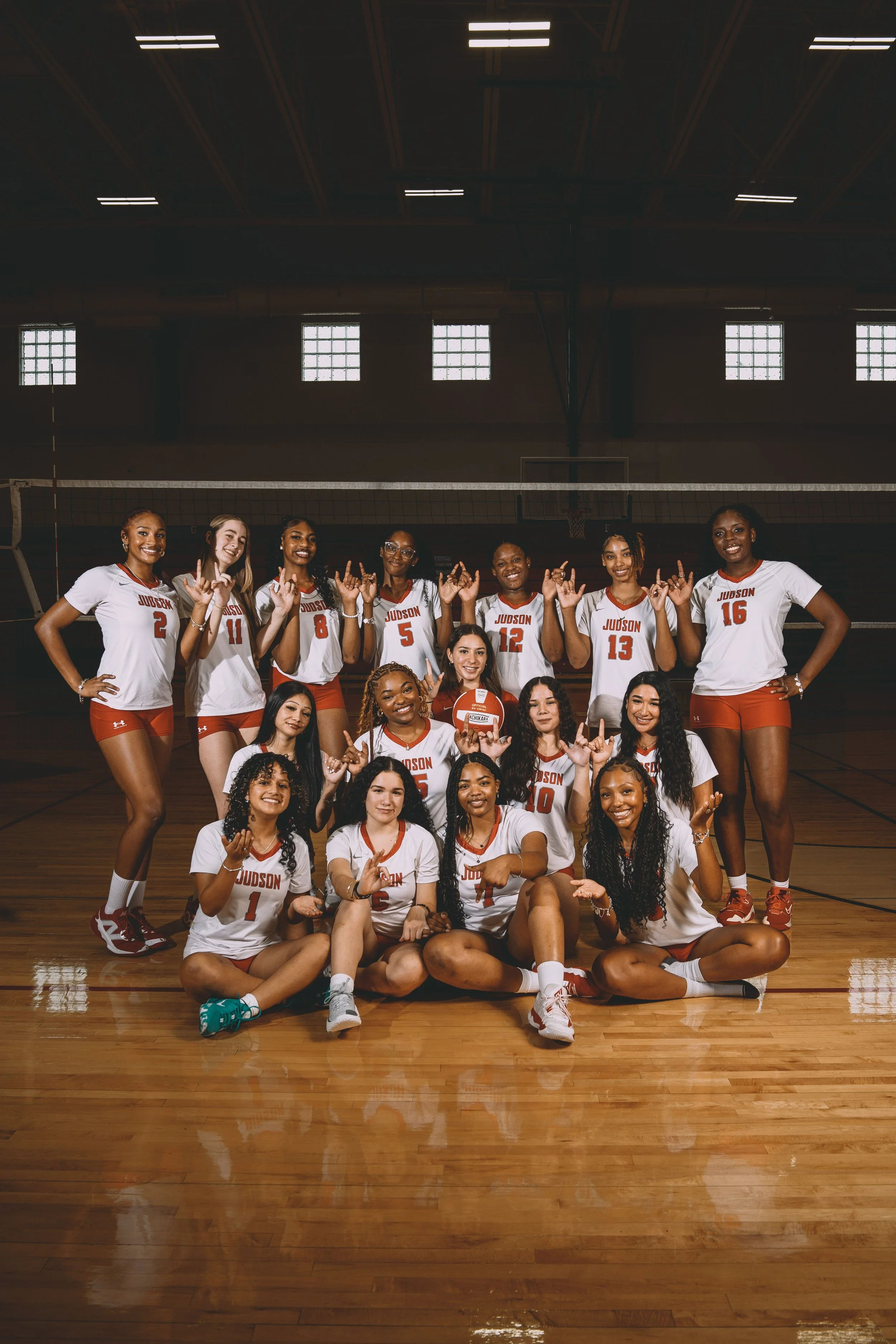 A women's volleyball team in a gymnasium, wearing white and red jerseys, posing for a team photo.