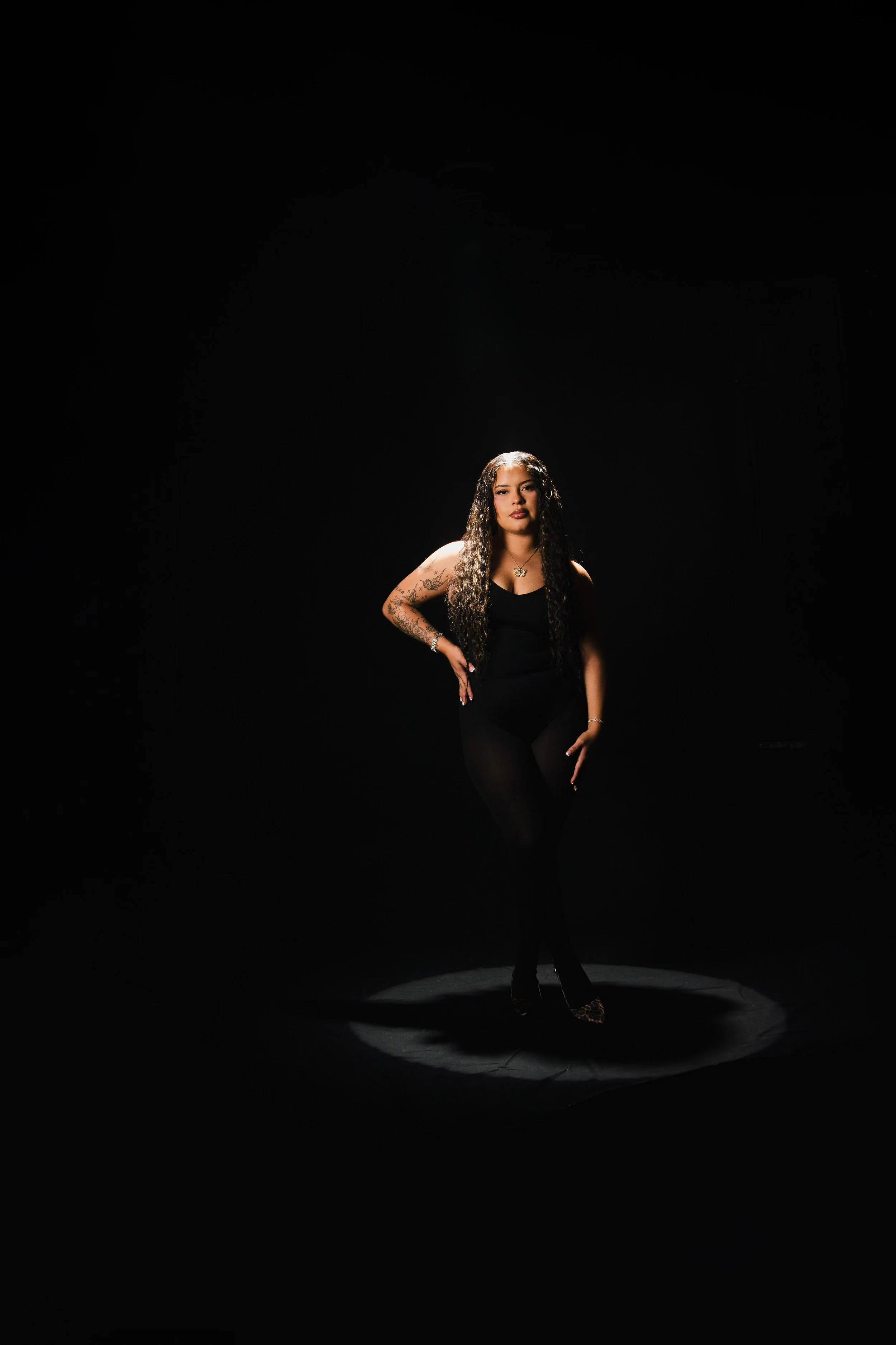 A woman with long curly hair, wearing a black outfit, standing in a spotlight on a dark background.