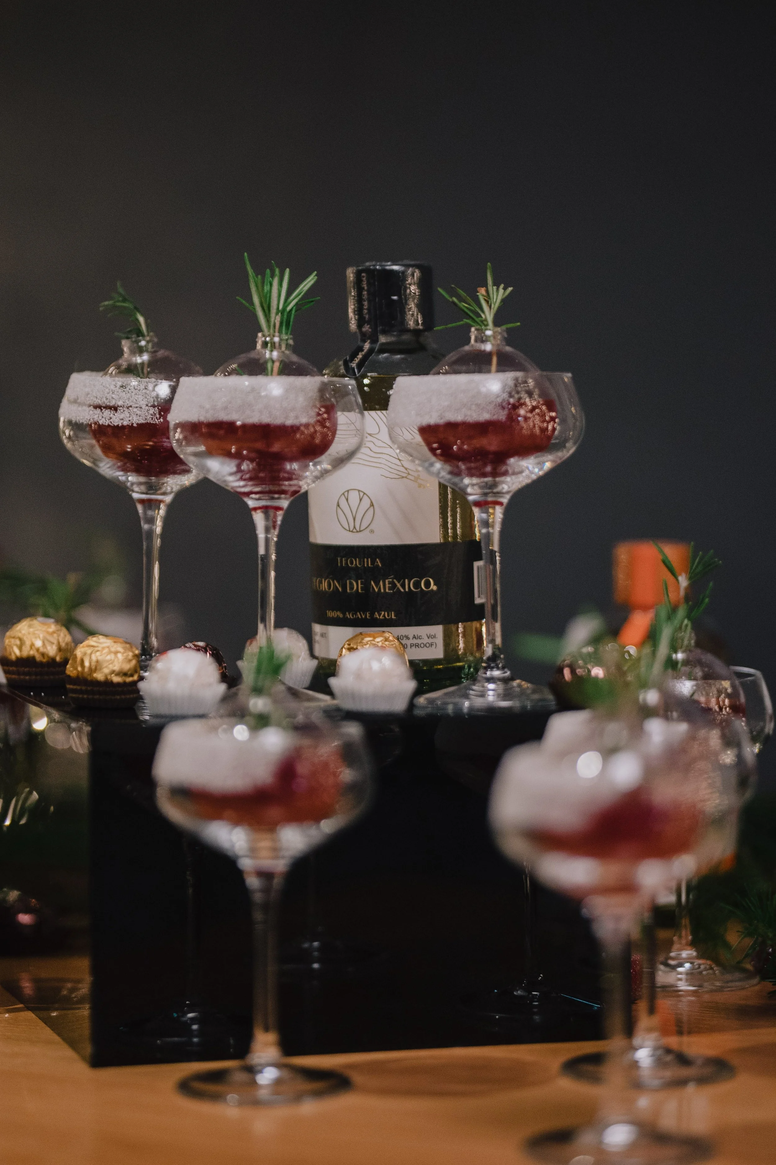 A bottle of tequila surrounded by several coupe glasses filled with red-colored drinks, garnished with small sprigs of rosemary.