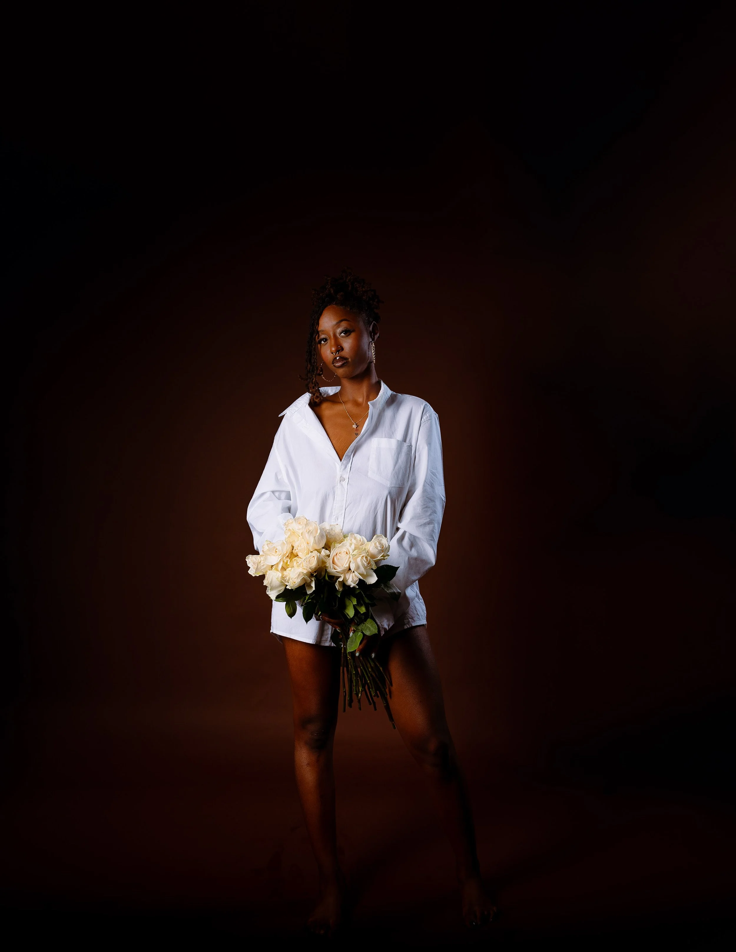 A woman standing against a dark background, holding a bouquet of white roses, wearing an oversized white shirt, and looking at the camera.