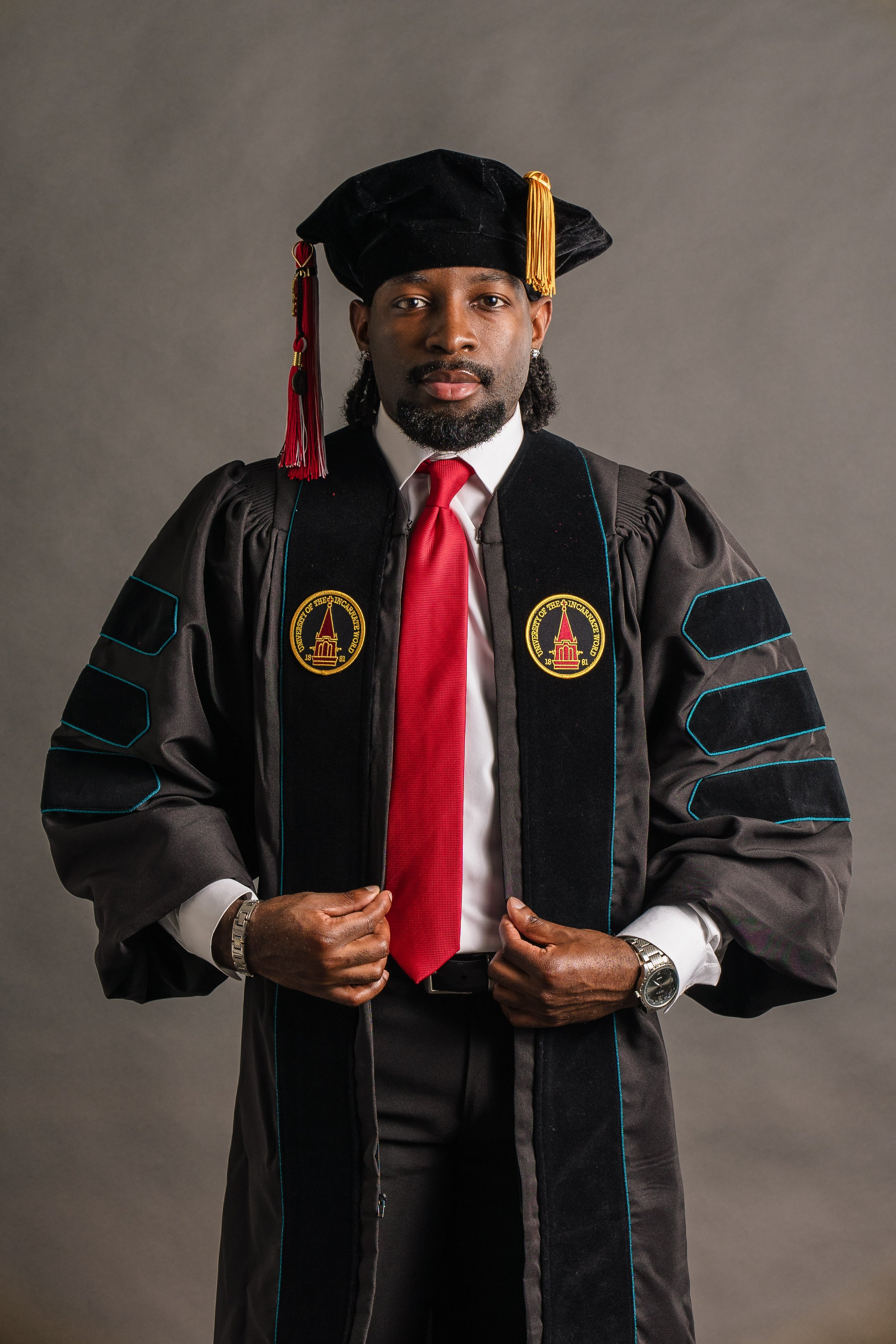 A man in graduation regalia, wearing a black cap with gold and red tassels and an academic gown with patches on the sleeves. He has a white shirt, red tie, and watches on his wrists, standing against a plain gray background.