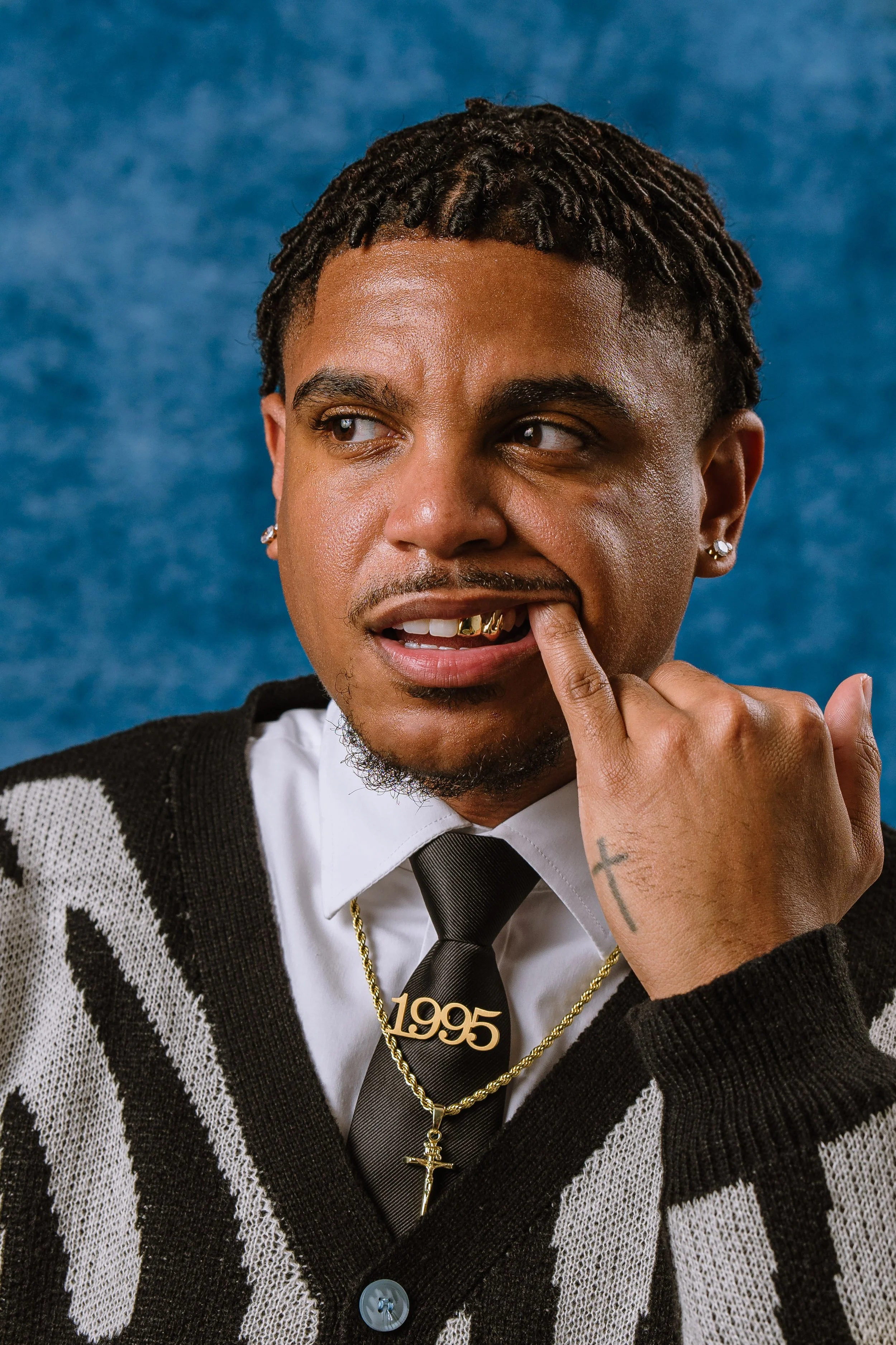 A man with dreadlocks, wearing a black and white sweater, white shirt, black tie, and gold jewelry, touches his lips with his finger, showing a gold grill and a tattoo of a cross on his hand. The background is blue.
